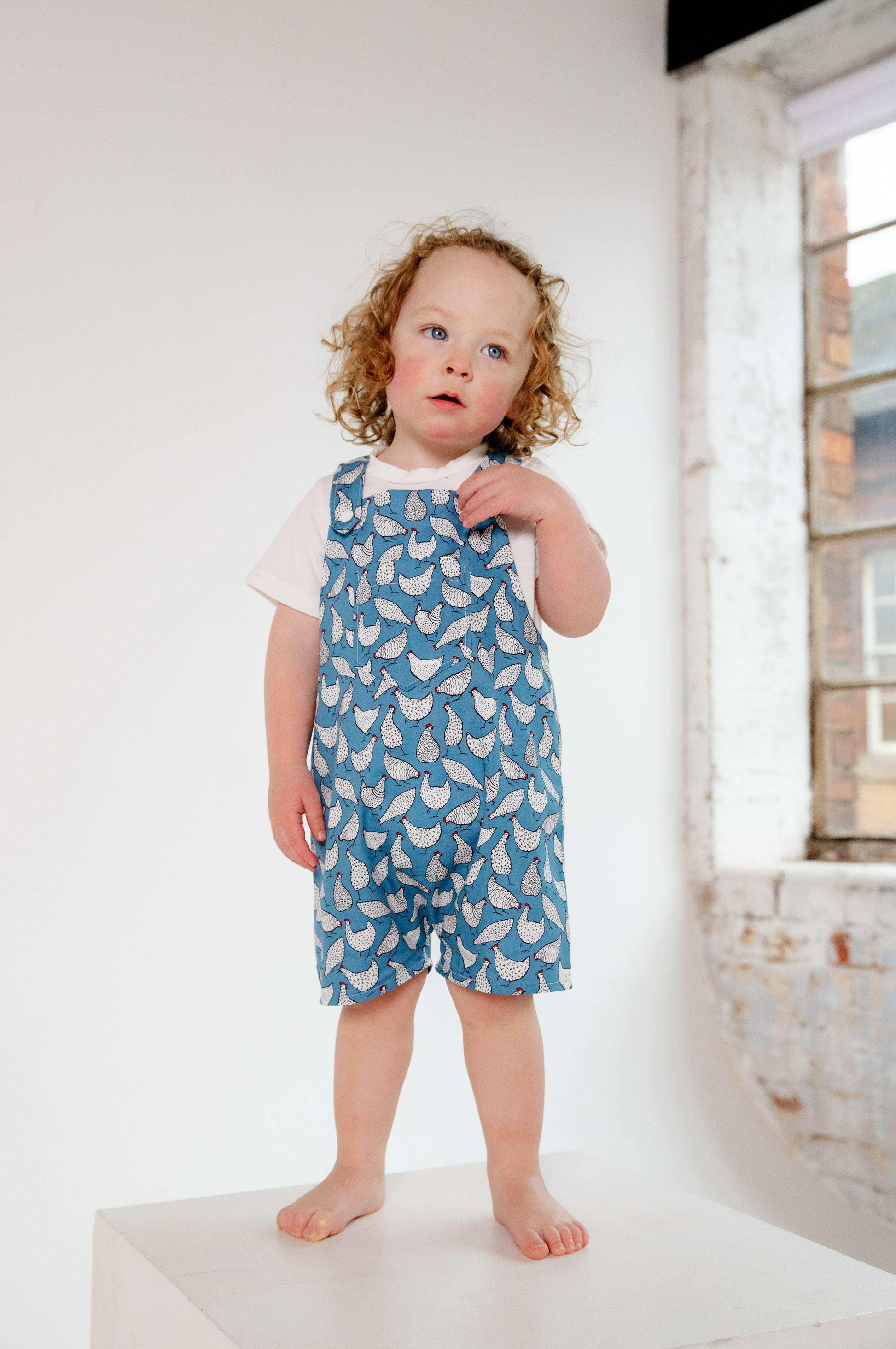 A toddler wearing a handmade blue and white chicken romper crafted from cotton fabric. The romper features, a front pocket, and a loose comfortable fit. The outfit is a timeless, classic design, perfect for all occasions.