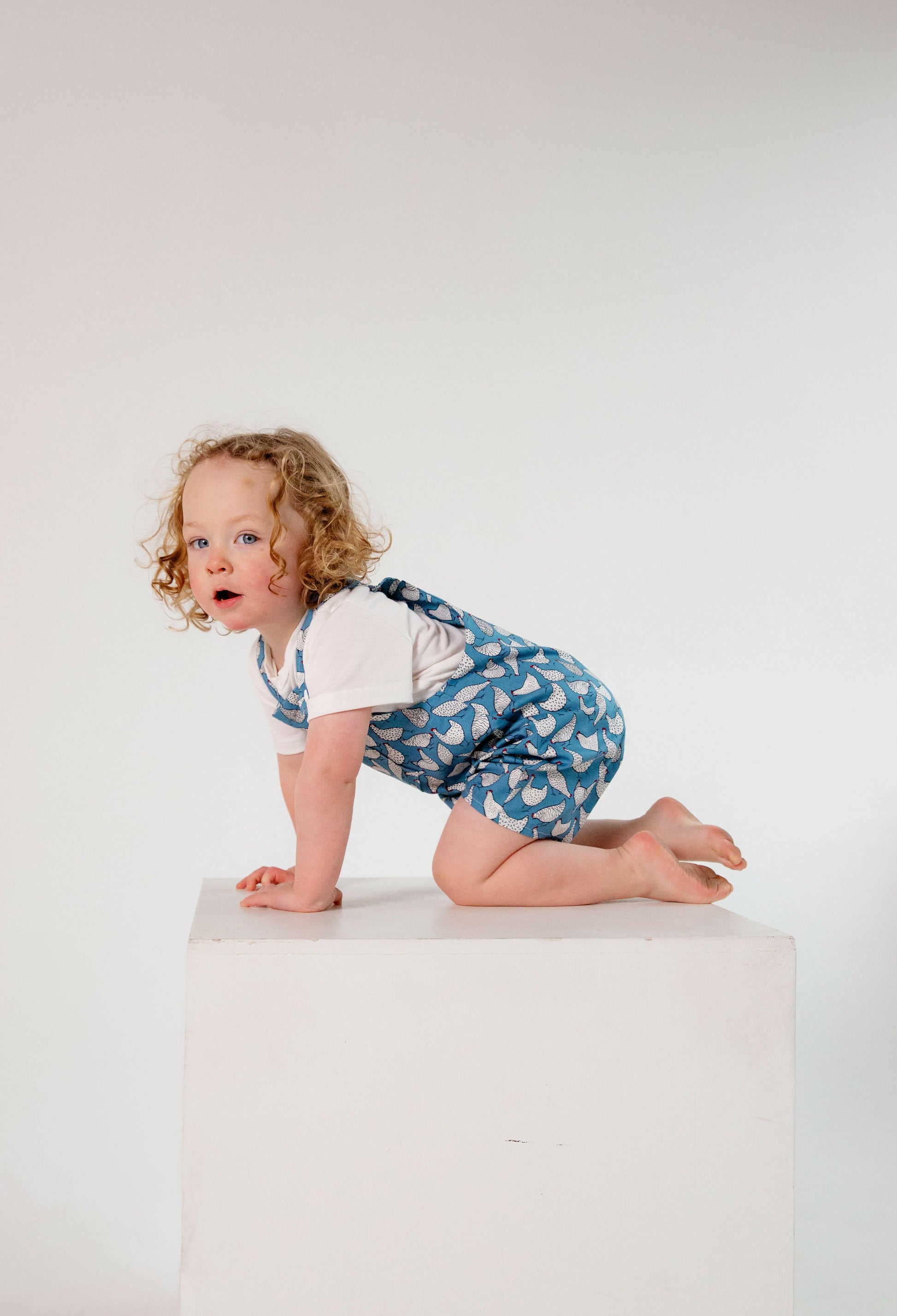 A toddler wearing a handmade blue and white chicken romper crafted from cotton fabric. The romper features, a front pocket, and a loose comfortable fit. The outfit is a timeless, classic design, perfect for all occasions.
