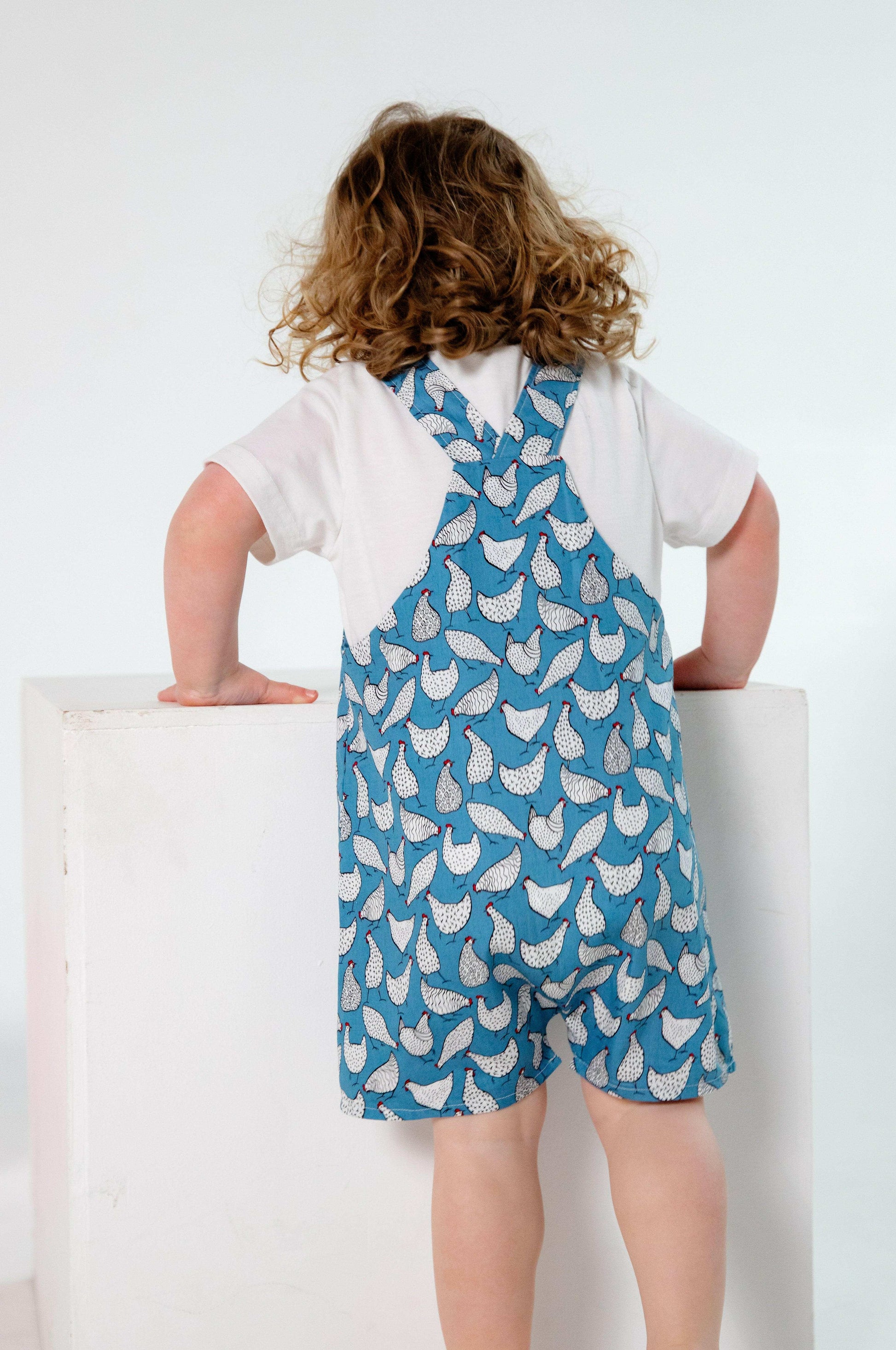 A toddler wearing a handmade blue and white chicken romper crafted from cotton fabric. The romper features, a front pocket, and a loose comfortable fit. The outfit is a timeless, classic design, perfect for all occasions.