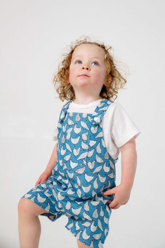 A toddler wearing a handmade blue and white chicken romper crafted from cotton fabric. The romper features, a front pocket, and a loose comfortable fit. The outfit is a timeless, classic design, perfect for all occasions.