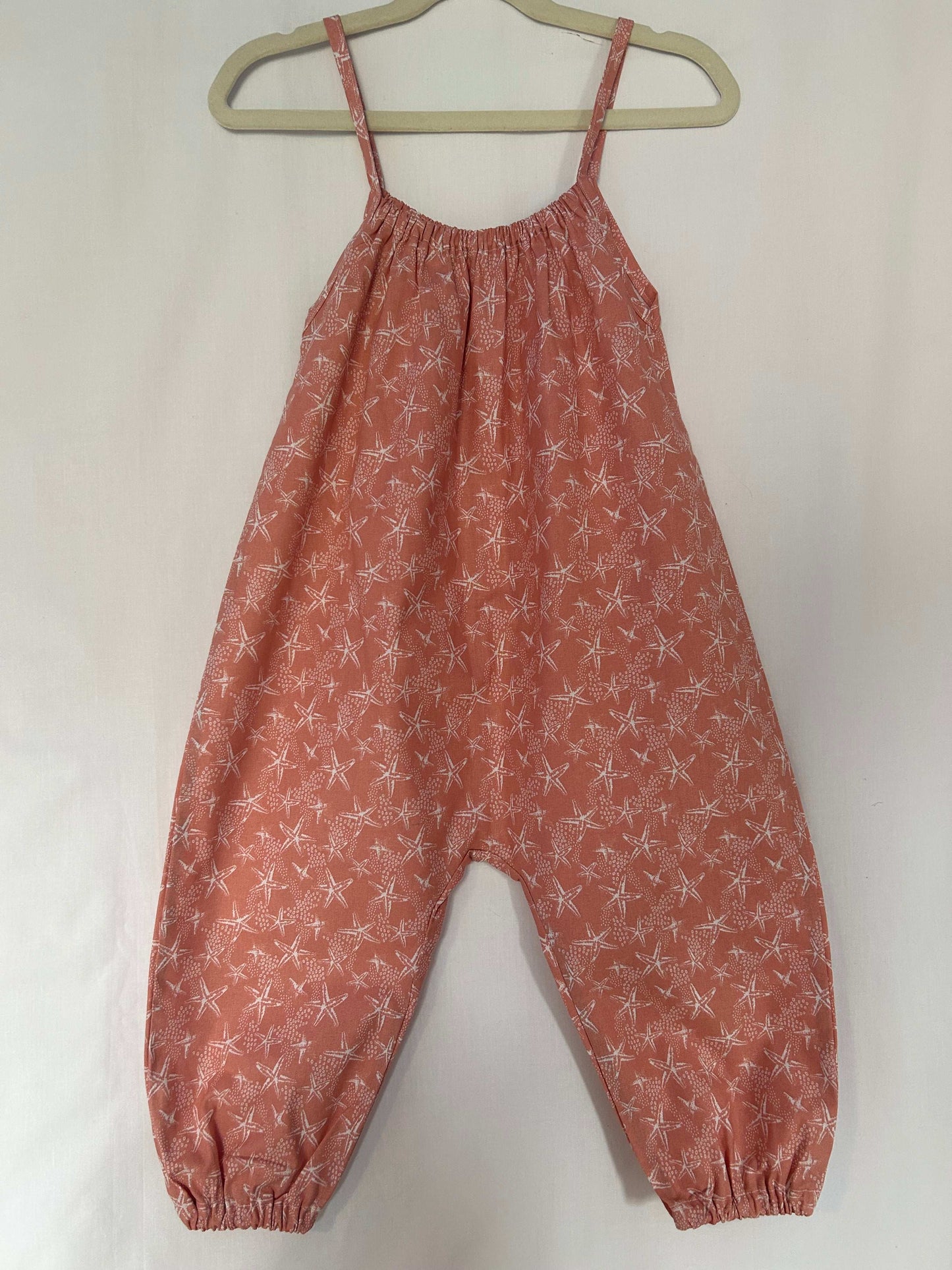 A young child wearing a handmade starfish print romper. The romper has a coral starfish print on soft, lightweight fabric. It has thin, shoulder straps and ruched neckline, relaxed, comfortable fit.