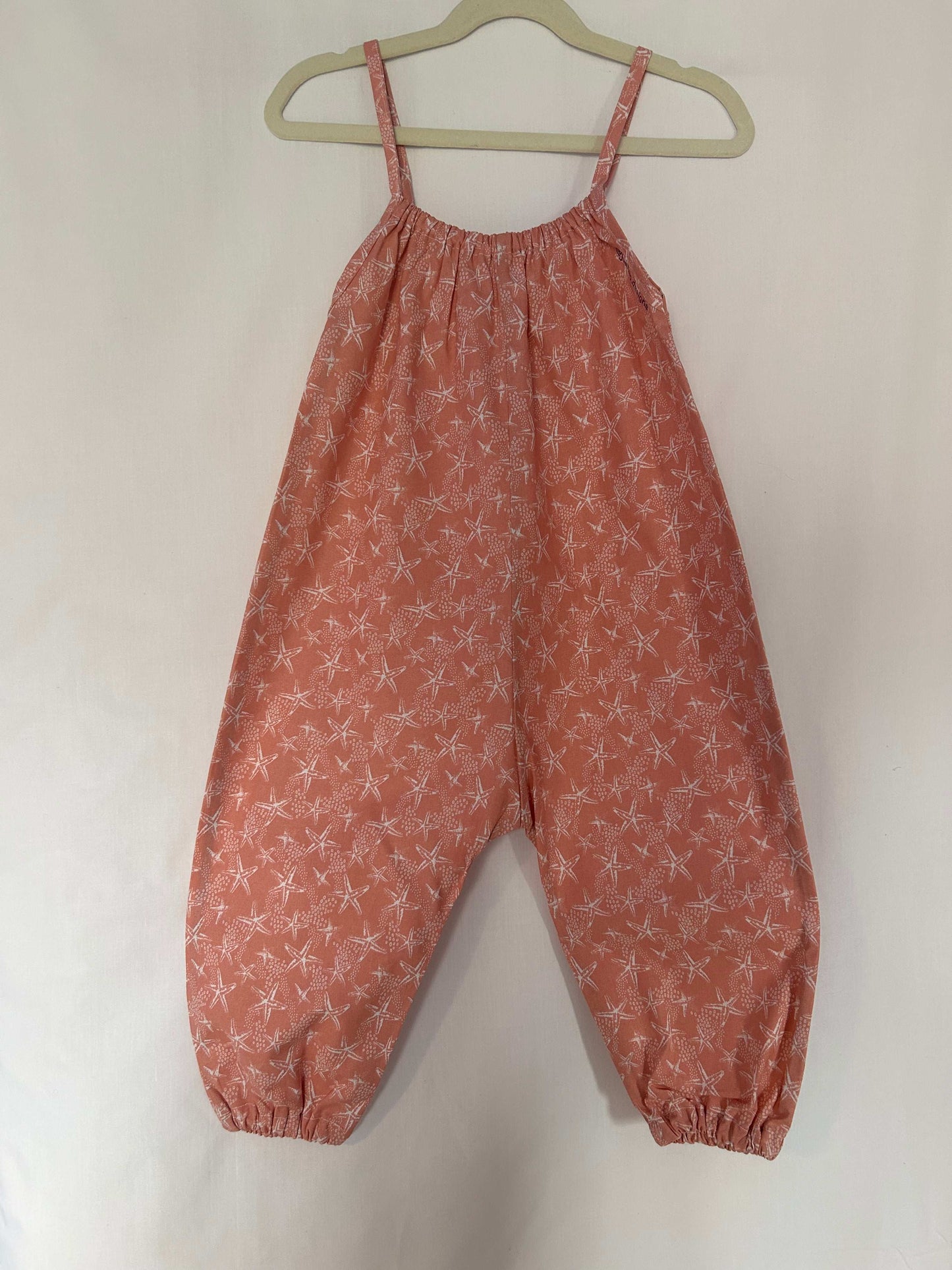 A young child wearing a handmade starfish print romper. The romper has a coral starfish print on soft, lightweight fabric. It has thin, shoulder straps and ruched neckline, relaxed, comfortable fit.