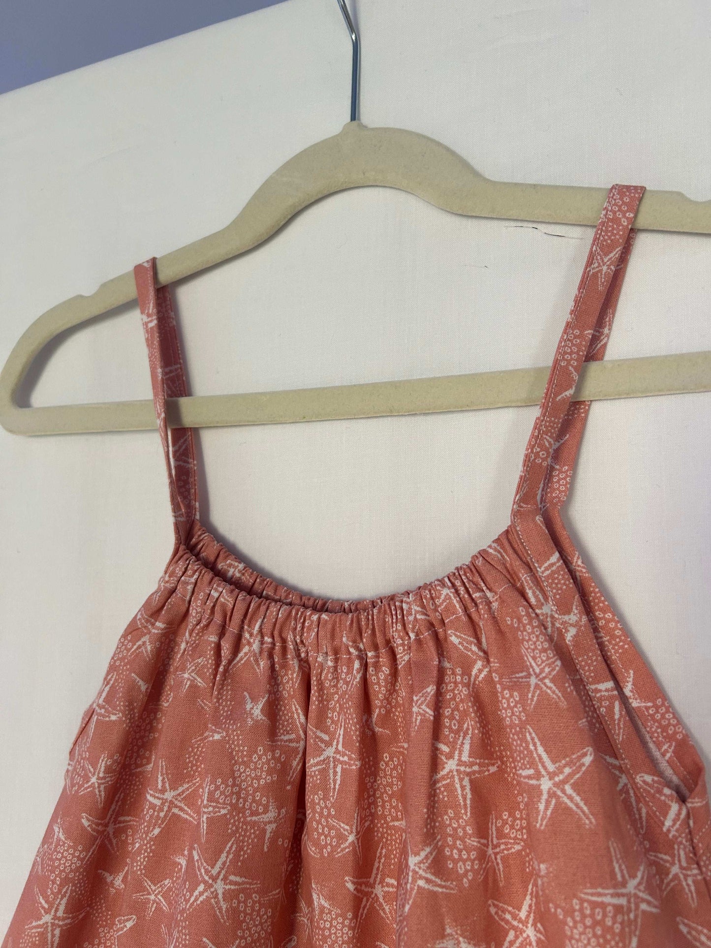 A young child wearing a handmade starfish print romper. The romper has a coral starfish print on soft, lightweight fabric. It has thin, shoulder straps and ruched neckline, relaxed, comfortable fit.