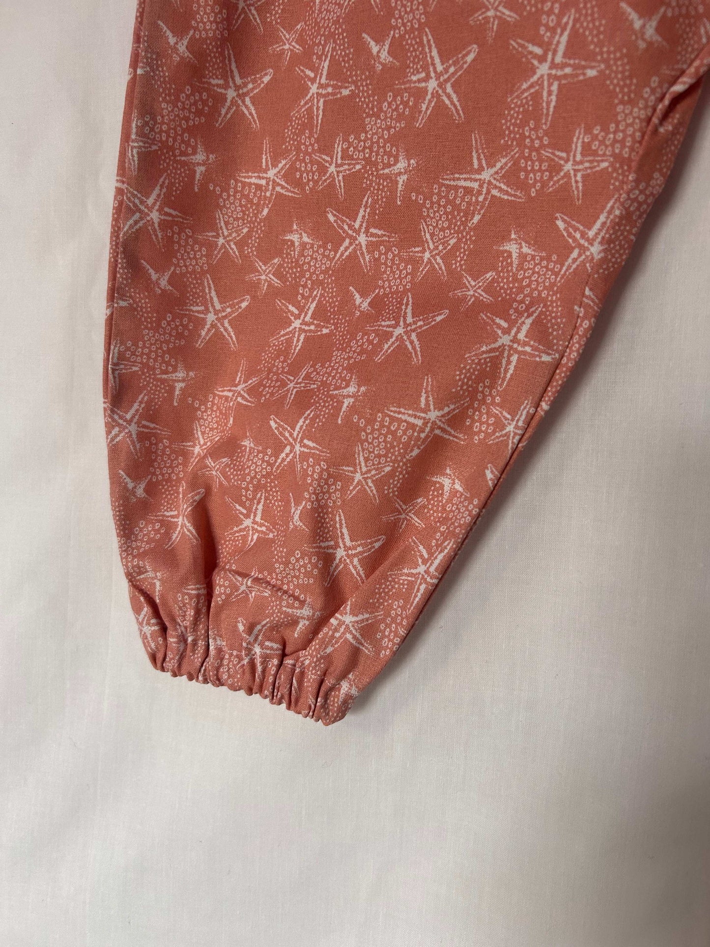 A young child wearing a handmade starfish print romper. The romper has a coral starfish print on soft, lightweight fabric. It has thin, shoulder straps and ruched neckline, relaxed, comfortable fit.