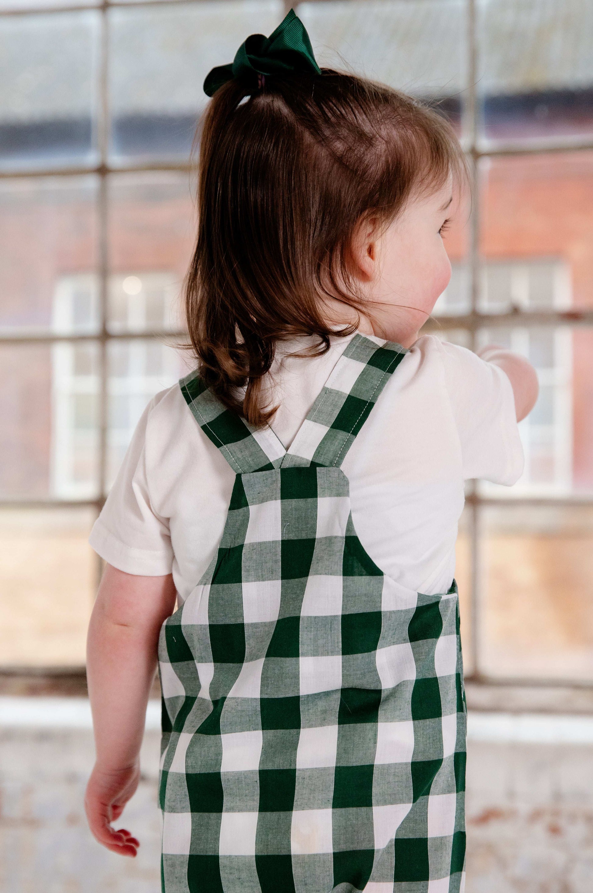 A toddler wearing a handmade green and white gingham romper crafted from cotton fabric. The romper features, a front pocket, and a loose comfortable fit. The outfit is a timeless, classic design, perfect for all occasions.