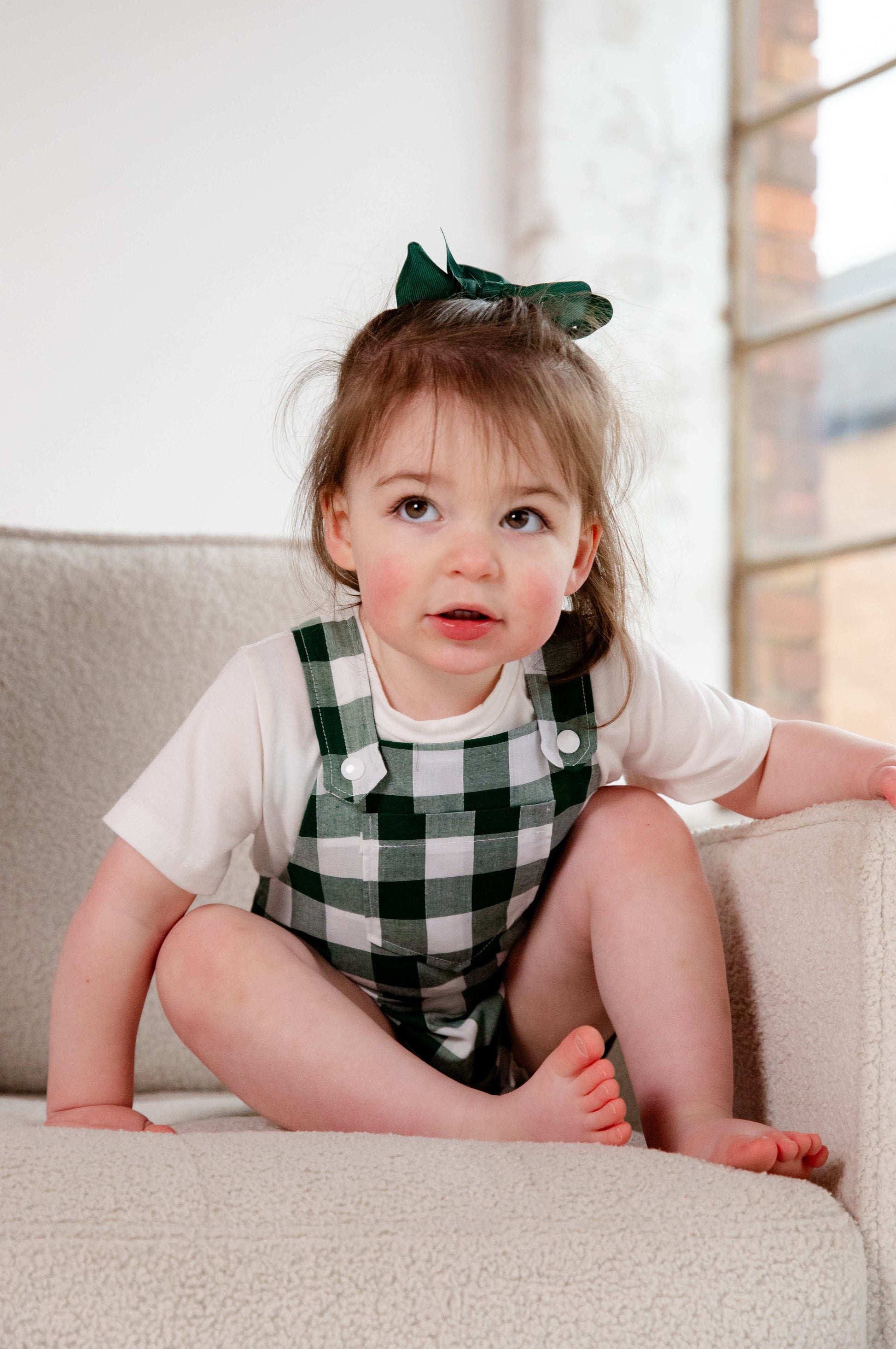 A toddler wearing a handmade green and white gingham romper crafted from cotton fabric. The romper features, a front pocket, and a loose comfortable fit. The outfit is a timeless, classic design, perfect for all occasions.