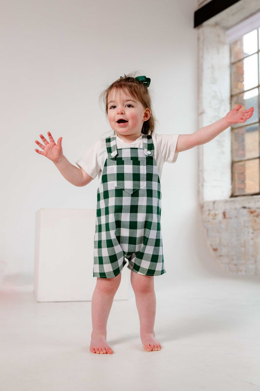 A toddler wearing a handmade green and white gingham romper crafted from cotton fabric. The romper features, a front pocket, and a loose comfortable fit. The outfit is a timeless, classic design, perfect for all occasions.