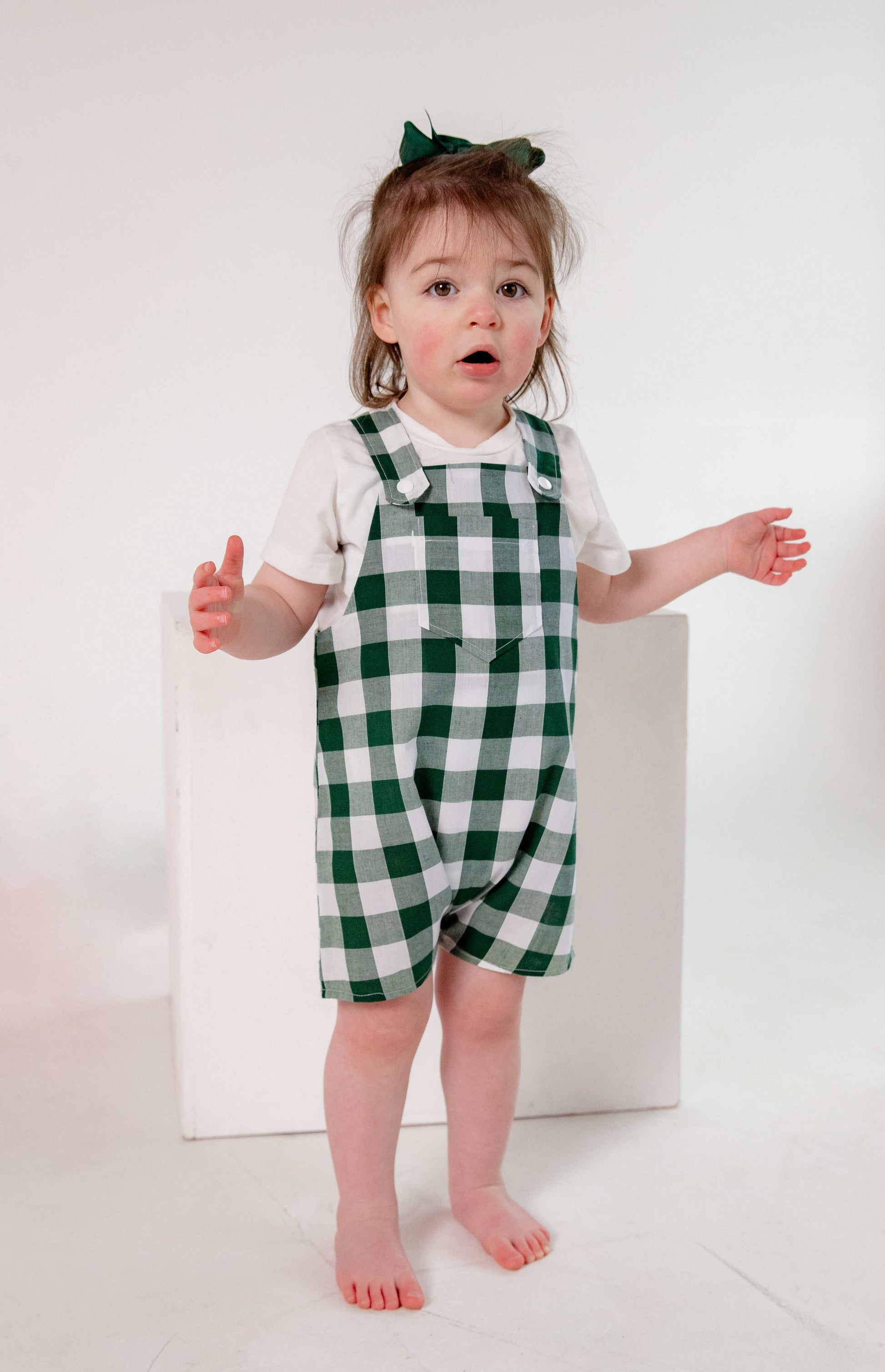 A toddler wearing a handmade green and white gingham romper crafted from cotton fabric. The romper features, a front pocket, and a loose comfortable fit. The outfit is a timeless, classic design, perfect for all occasions.