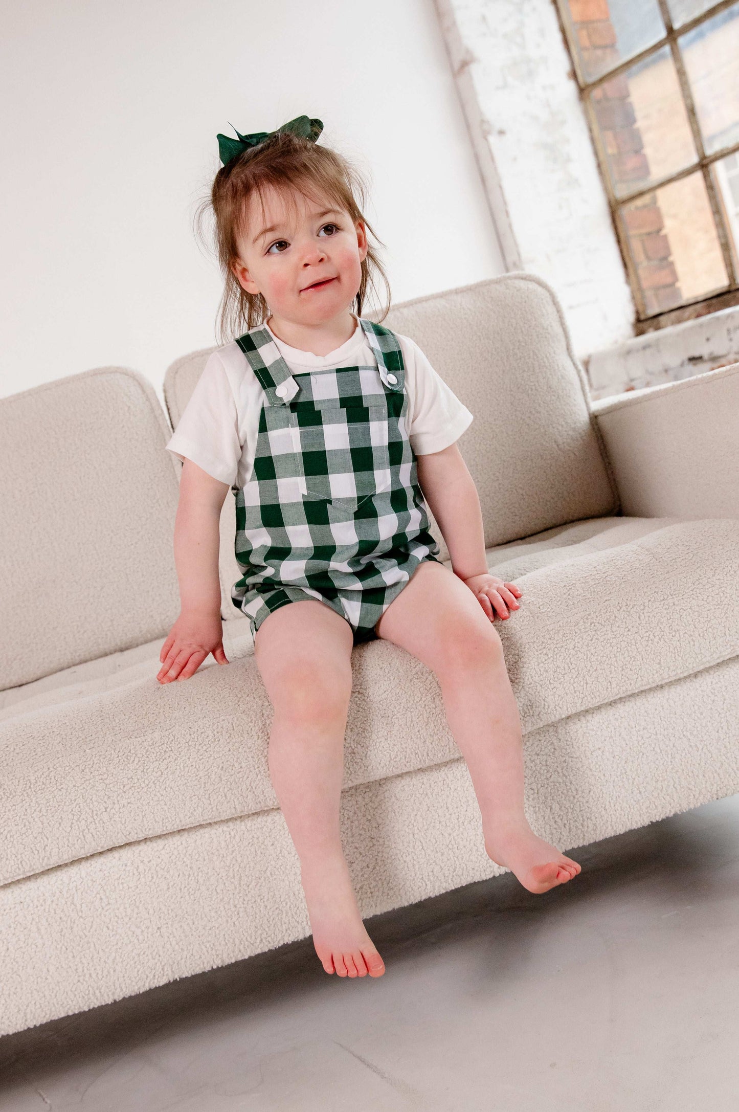 A toddler wearing a handmade green and white gingham romper crafted from cotton fabric. The romper features, a front pocket, and a loose comfortable fit. The outfit is a timeless, classic design, perfect for all occasions.