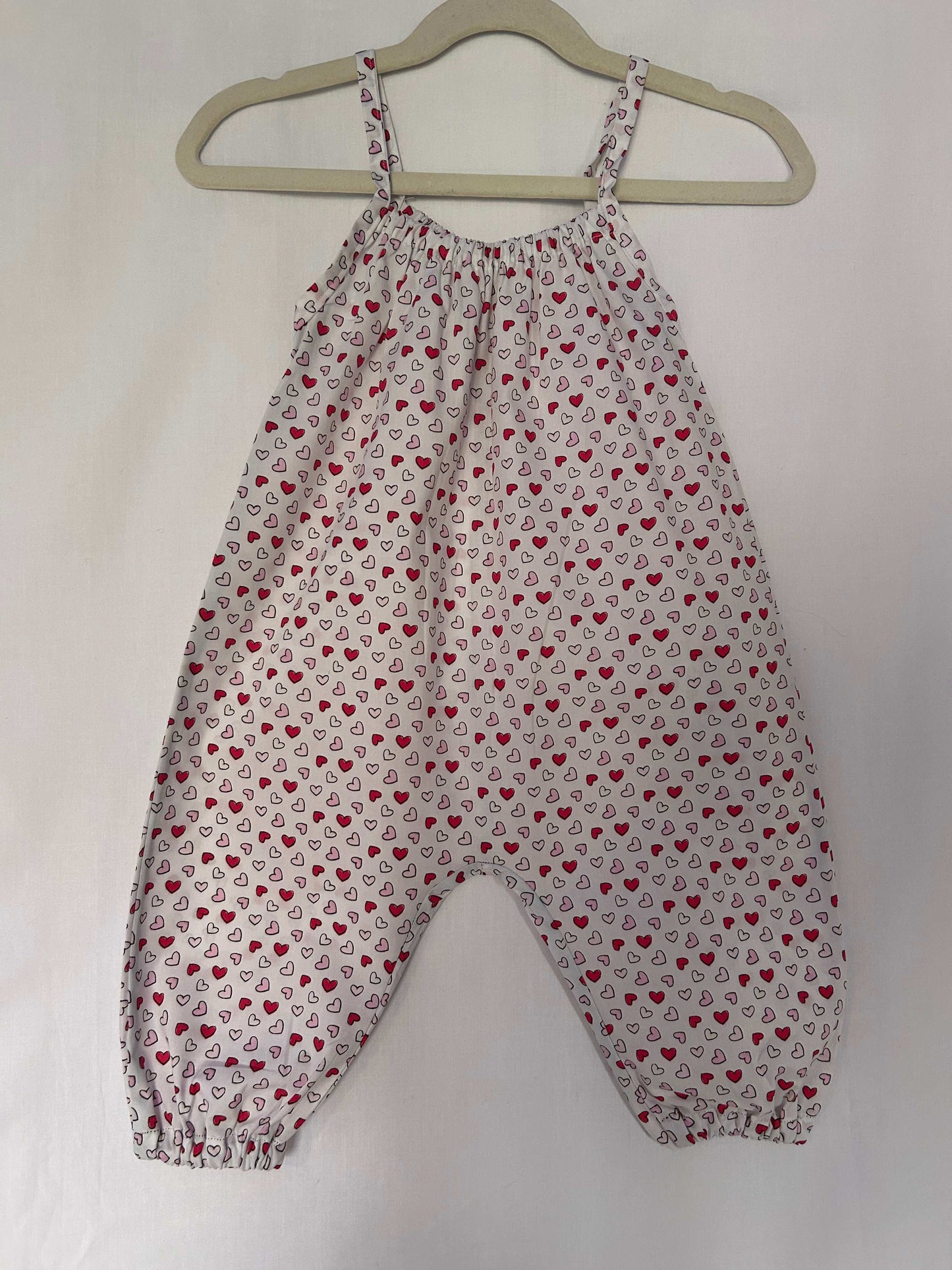 A young child wearing a handmade heart print romper. The romper has a bold heart print on soft, lightweight fabric. It has thin, shoulder straps and ruched neckline, relaxed, comfortable fit.