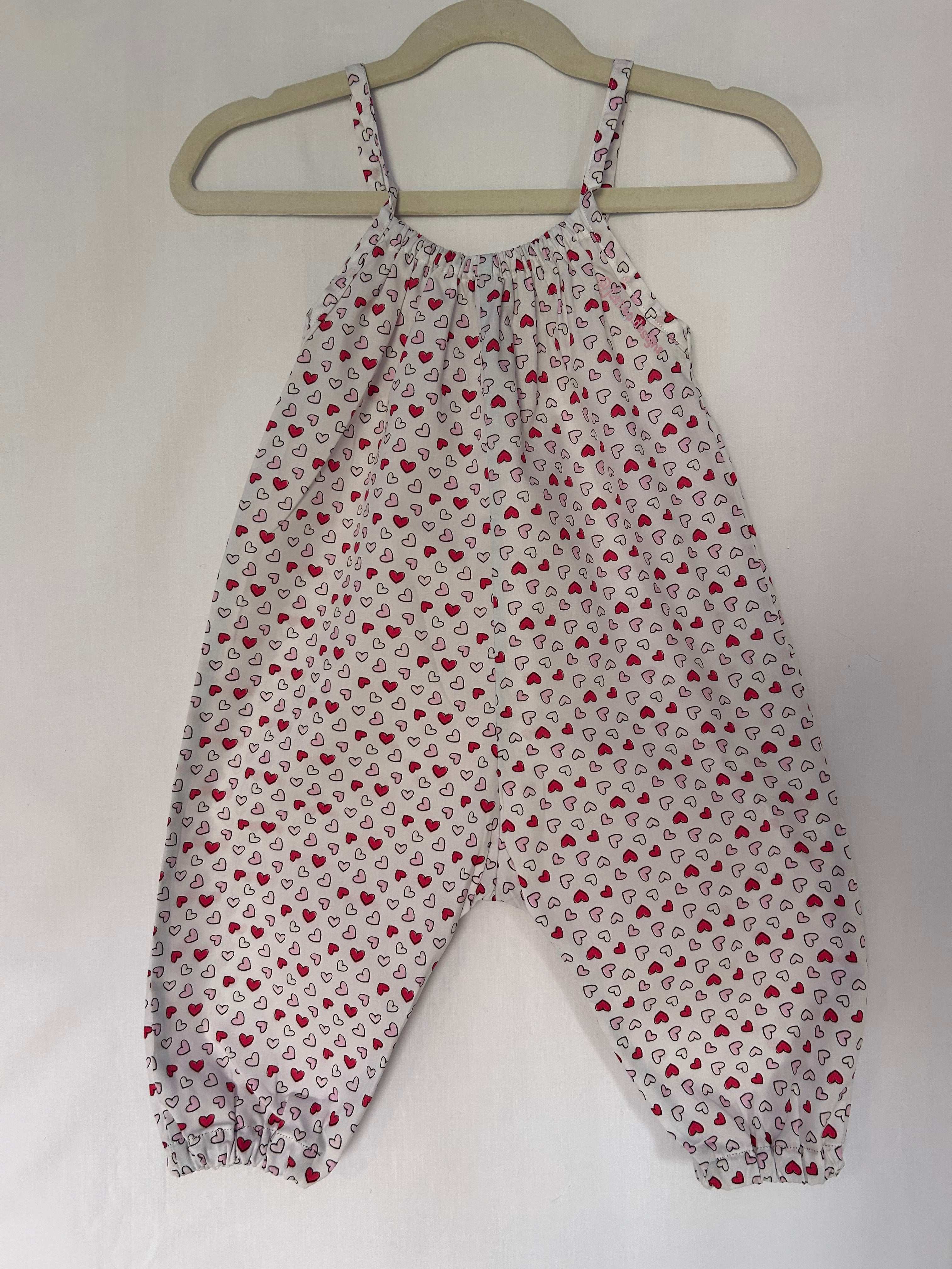 A young child wearing a handmade heart print romper. The romper has a bold heart print on soft, lightweight fabric. It has thin, shoulder straps and ruched neckline, relaxed, comfortable fit.