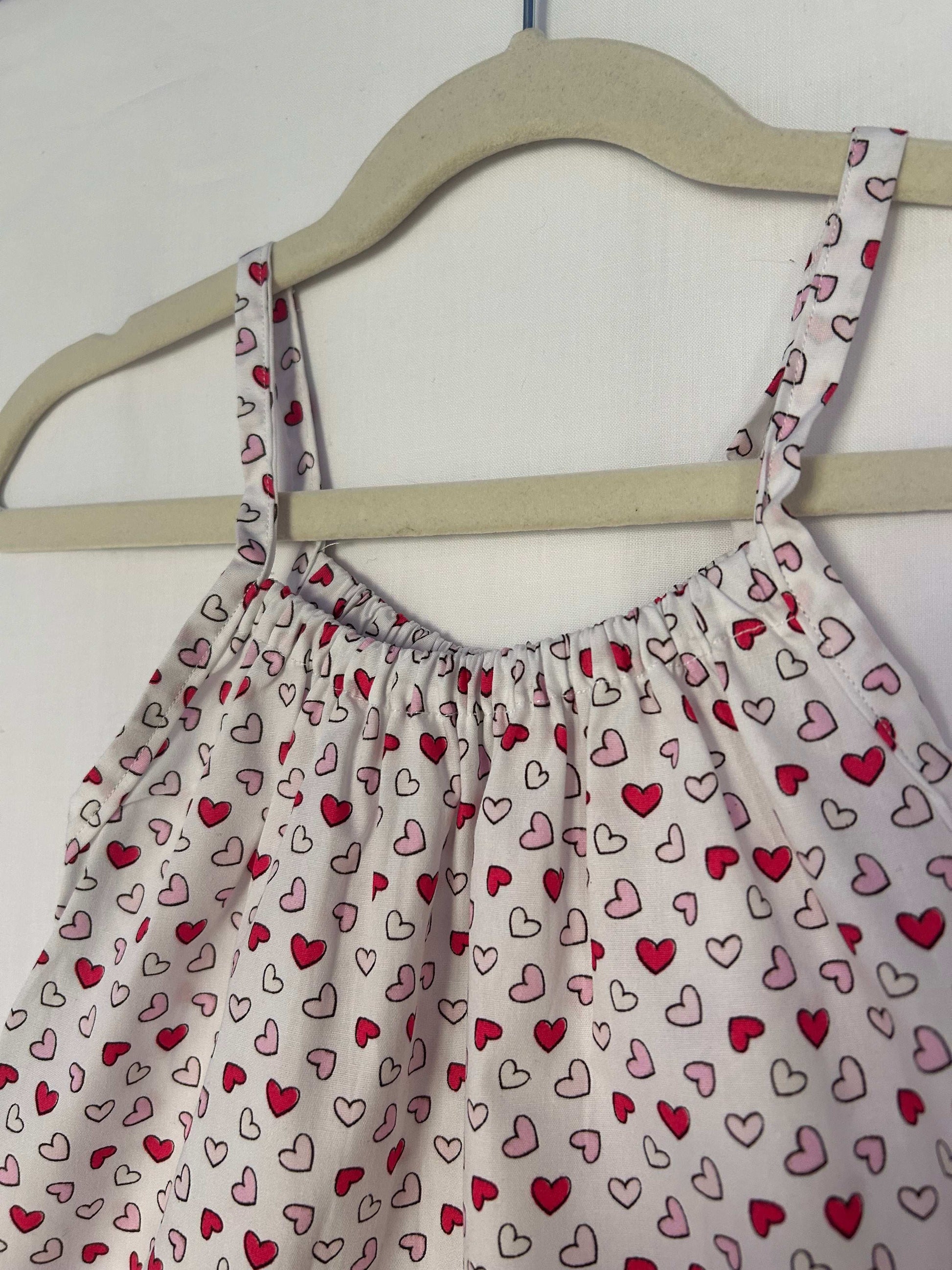 A young child wearing a handmade heart print romper. The romper has a bold heart print on soft, lightweight fabric. It has thin, shoulder straps and ruched neckline, relaxed, comfortable fit.