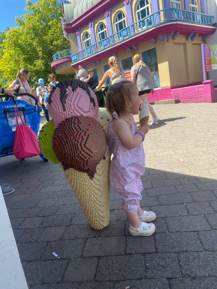 A young child wearing a handmade floral print romper. The romper has a lilac flower print on soft, lightweight fabric. It has thin, shoulder straps and ruched neckline, relaxed, comfortable fit.