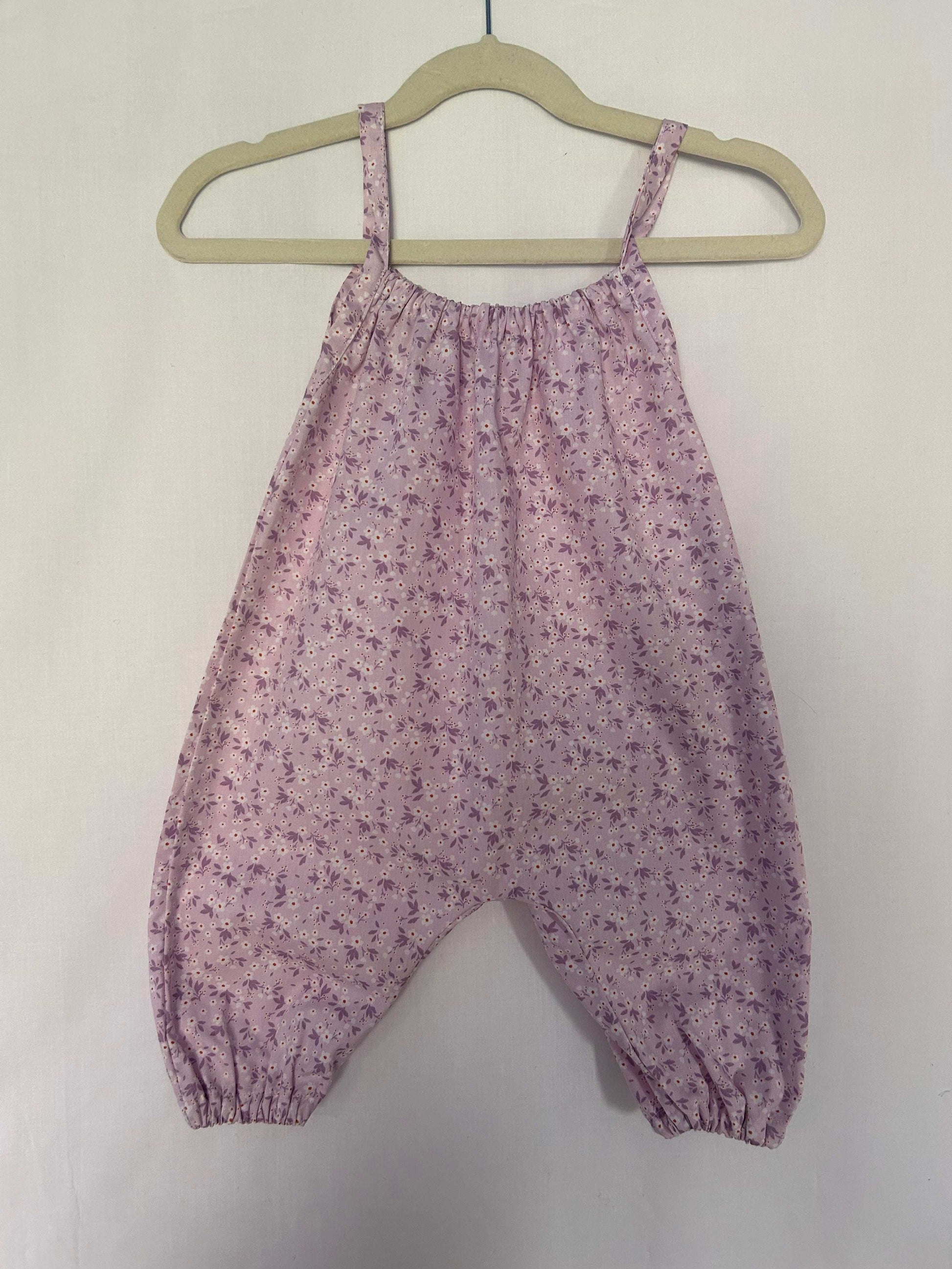A young child wearing a handmade floral print romper. The romper has a lilac flower print on soft, lightweight fabric. It has thin, shoulder straps and ruched neckline, relaxed, comfortable fit.