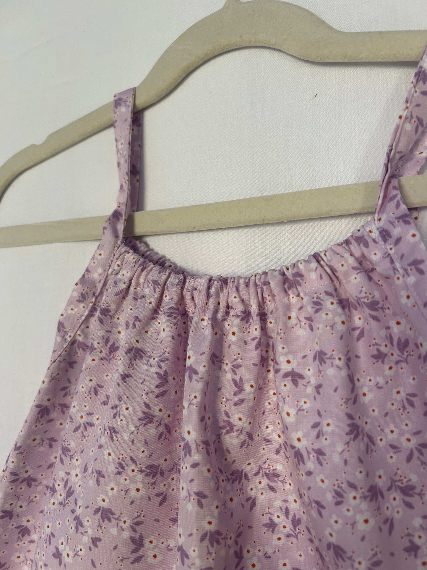 A young child wearing a handmade floral print romper. The romper has a lilac flower print on soft, lightweight fabric. It has thin, shoulder straps and ruched neckline, relaxed, comfortable fit.
