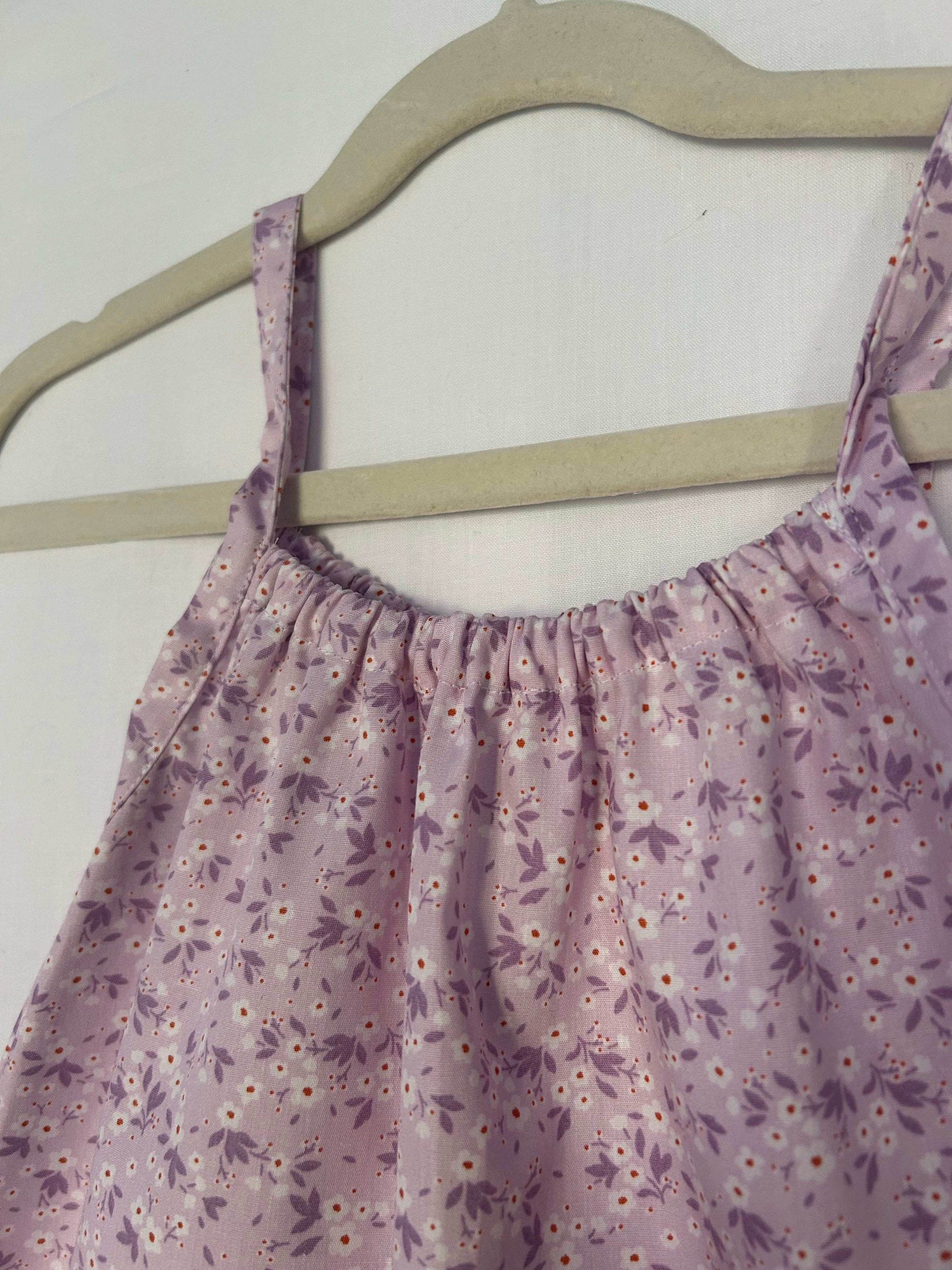 A young child wearing a handmade floral print romper. The romper has a lilac flower print on soft, lightweight fabric. It has thin, shoulder straps and ruched neckline, relaxed, comfortable fit.