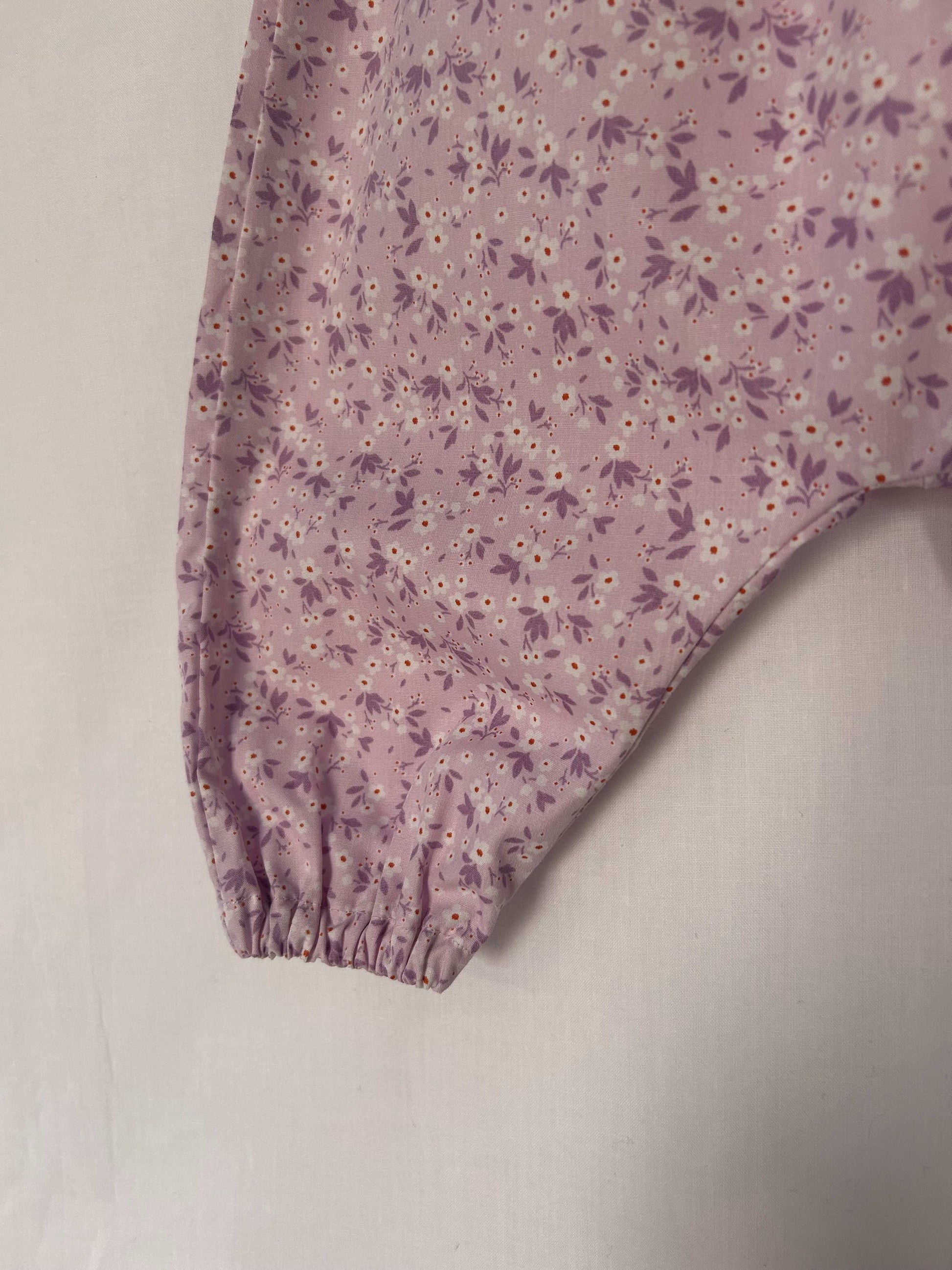 A young child wearing a handmade floral print romper. The romper has a lilac flower print on soft, lightweight fabric. It has thin, shoulder straps and ruched neckline, relaxed, comfortable fit.