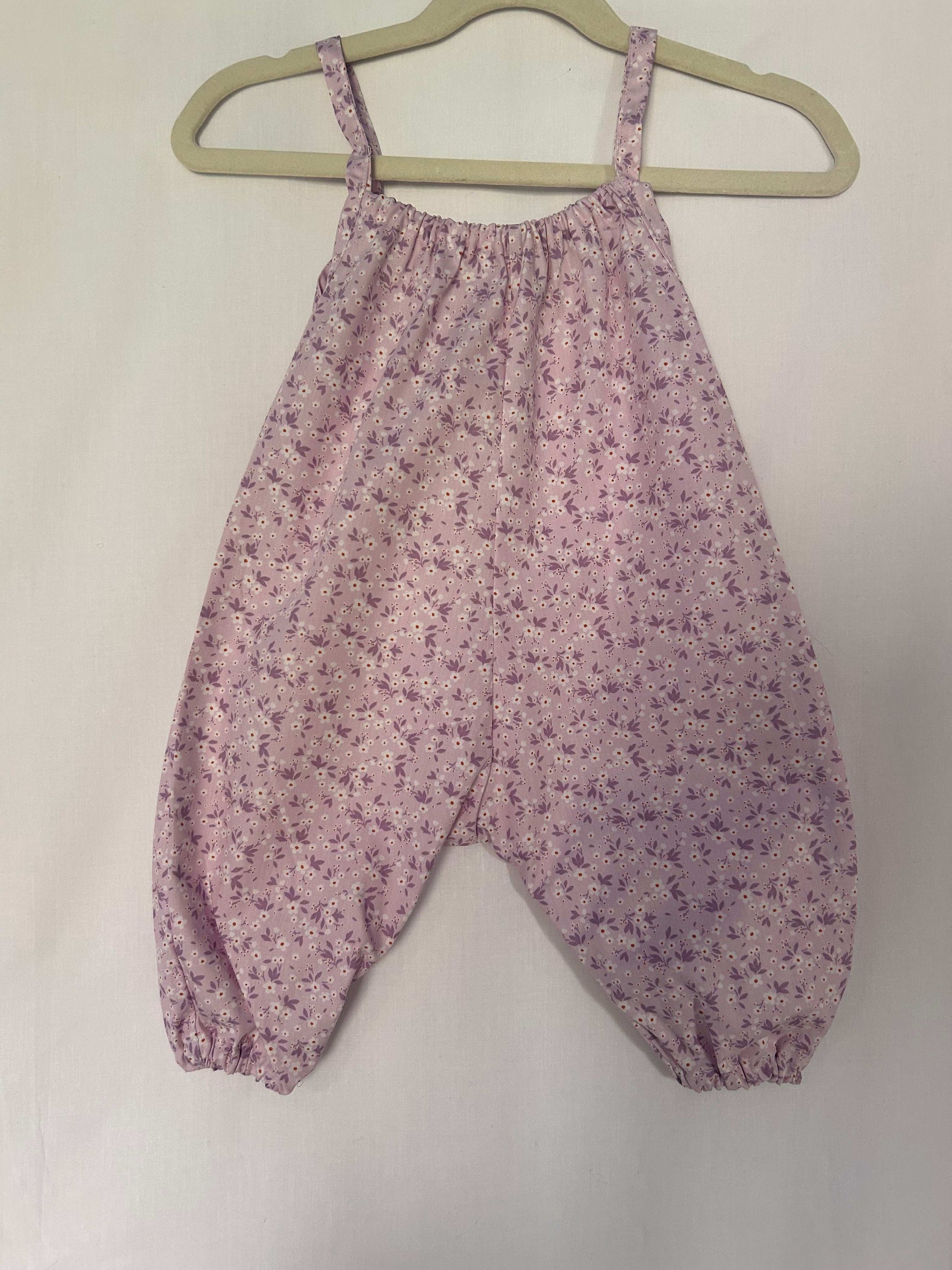 A young child wearing a handmade floral print romper. The romper has a lilac flower print on soft, lightweight fabric. It has thin, shoulder straps and ruched neckline, relaxed, comfortable fit.