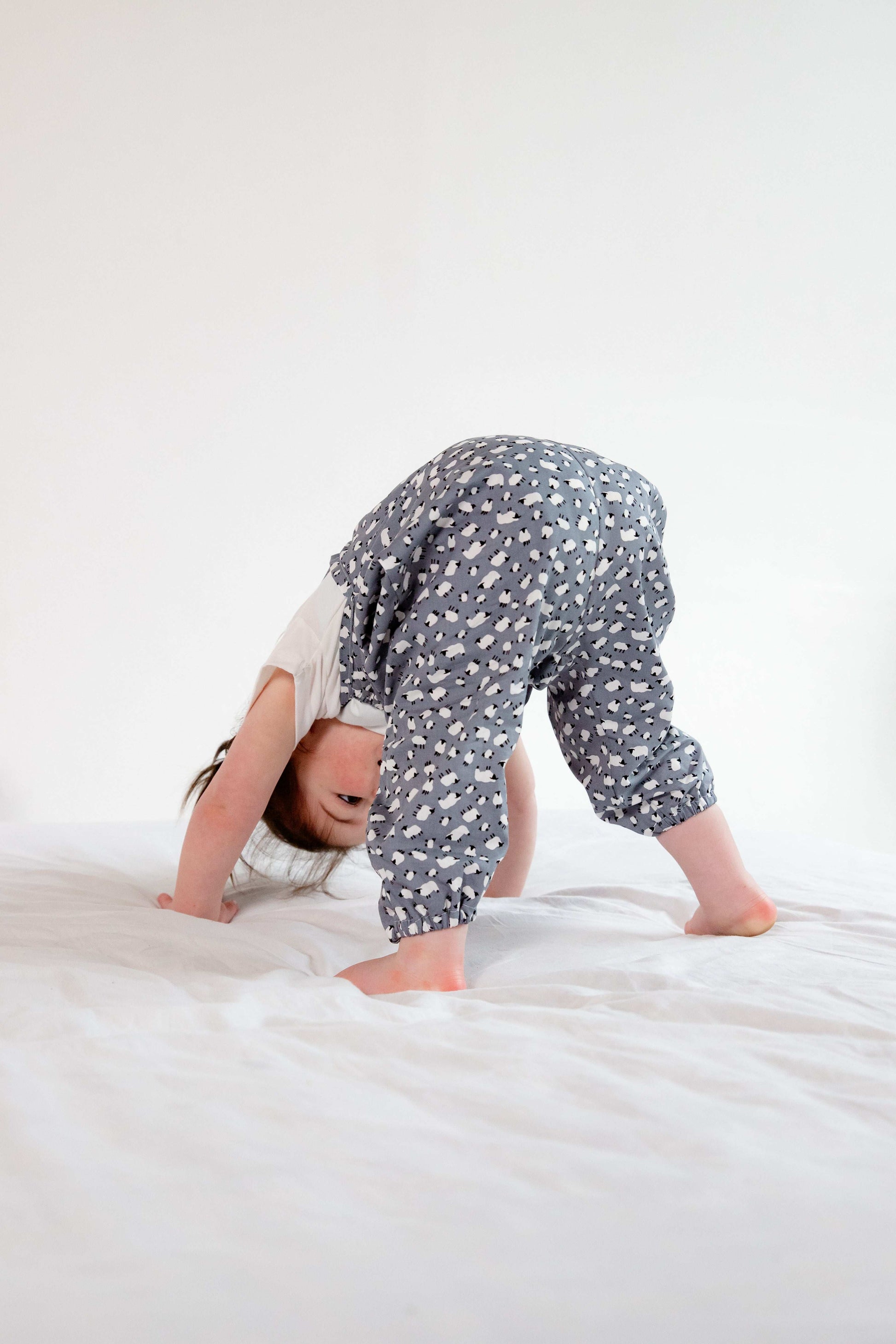 A young child wearing a handmade sheep romper. The romper has a delicate grey and white sheep print on soft, lightweight fabric. It has thin, shoulder straps and ruched neckline, relaxed, comfortable fit.