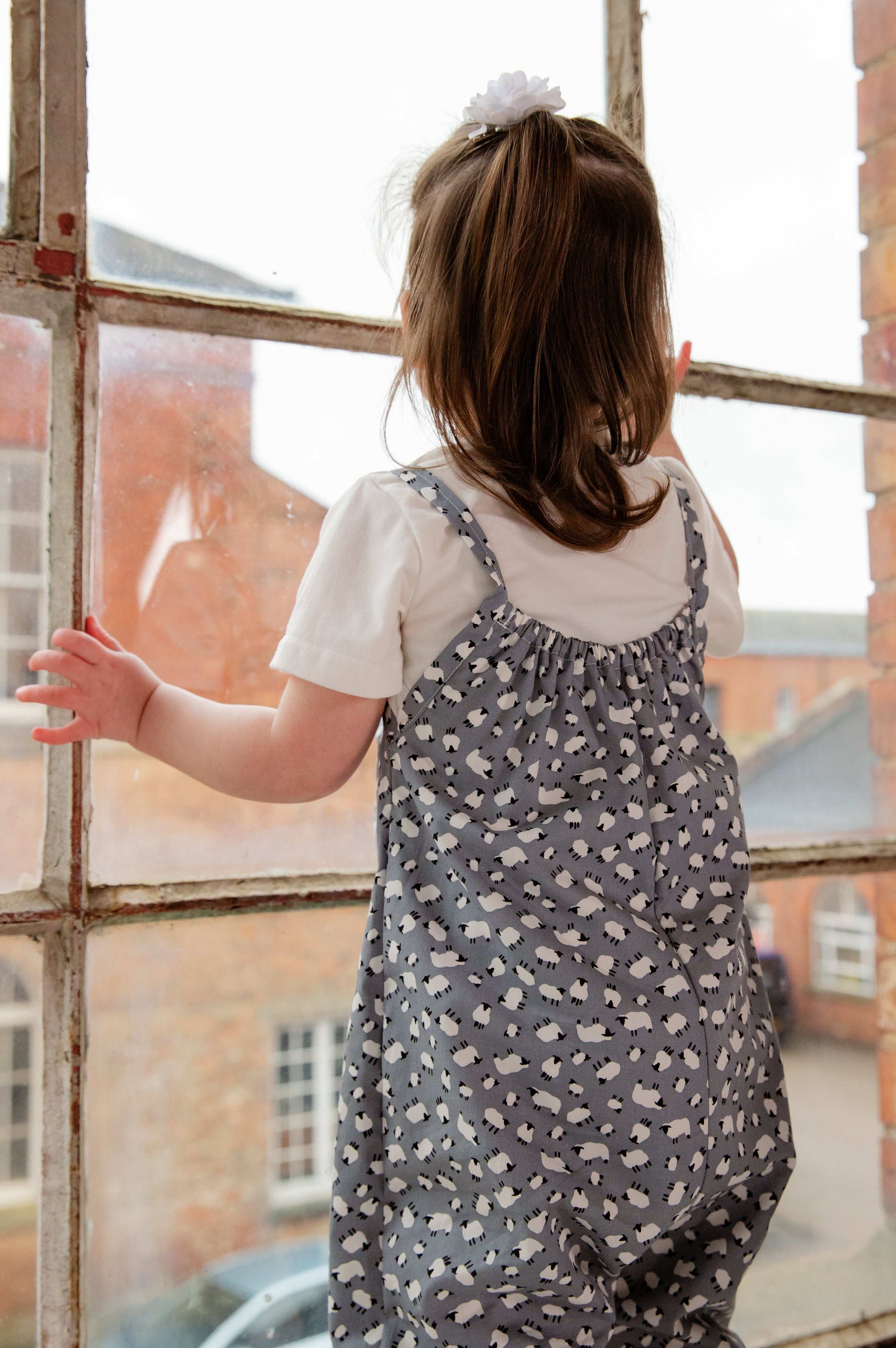 A young child wearing a handmade sheep romper. The romper has a delicate grey and white sheep print on soft, lightweight fabric. It has thin, shoulder straps and ruched neckline, relaxed, comfortable fit.