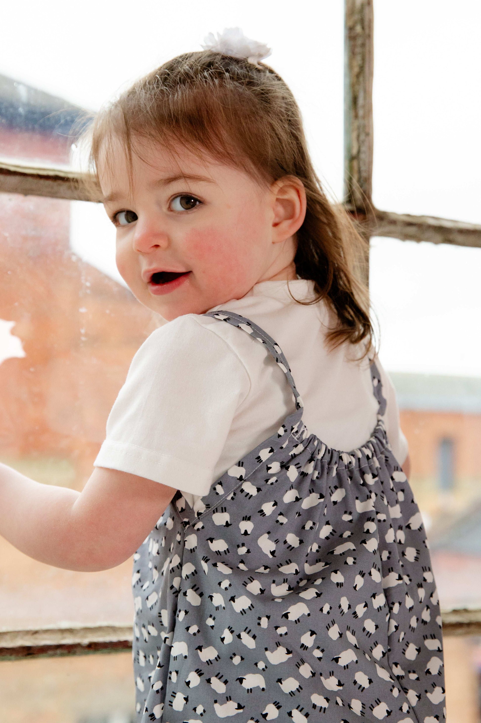 A young child wearing a handmade sheep romper. The romper has a delicate grey and white sheep print on soft, lightweight fabric. It has thin, shoulder straps and ruched neckline, relaxed, comfortable fit.