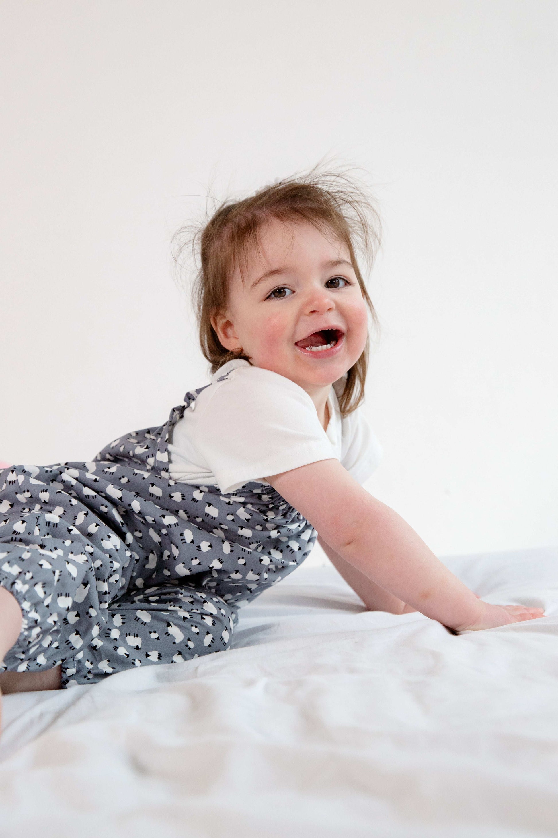 A young child wearing a handmade sheep romper. The romper has a delicate grey and white sheep print on soft, lightweight fabric. It has thin, shoulder straps and ruched neckline, relaxed, comfortable fit.