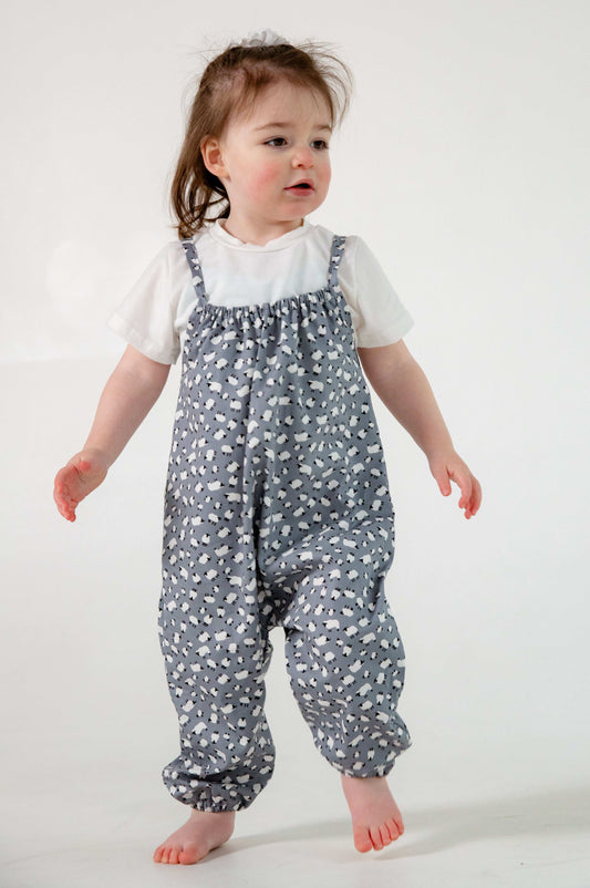 A young child wearing a handmade sheep romper. The romper has a delicate grey and white sheep print on soft, lightweight fabric. It has thin, shoulder straps and ruched neckline, relaxed, comfortable fit.