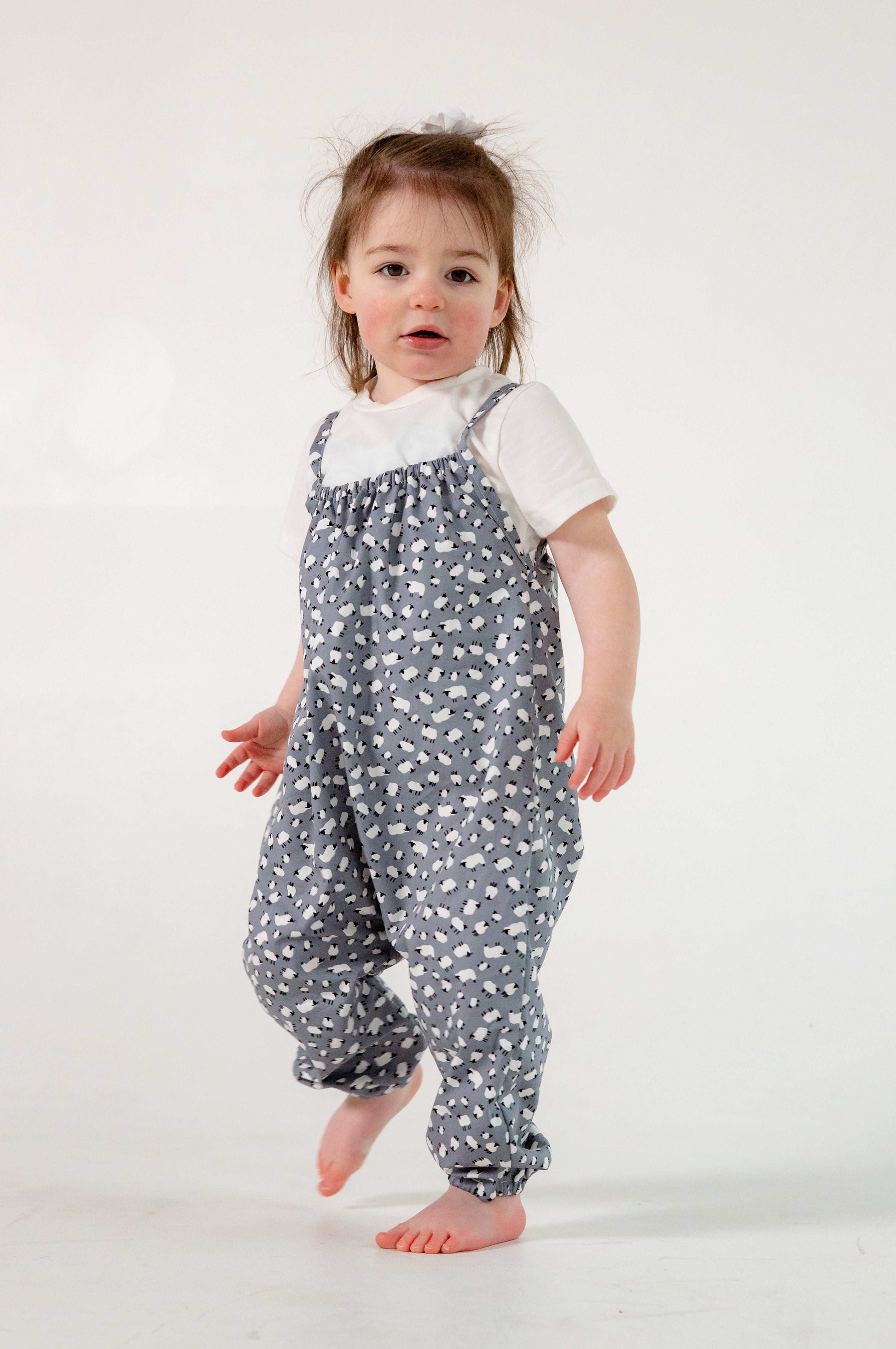 A young child wearing a handmade sheep romper. The romper has a delicate grey and white sheep print on soft, lightweight fabric. It has thin, shoulder straps and ruched neckline, relaxed, comfortable fit.
