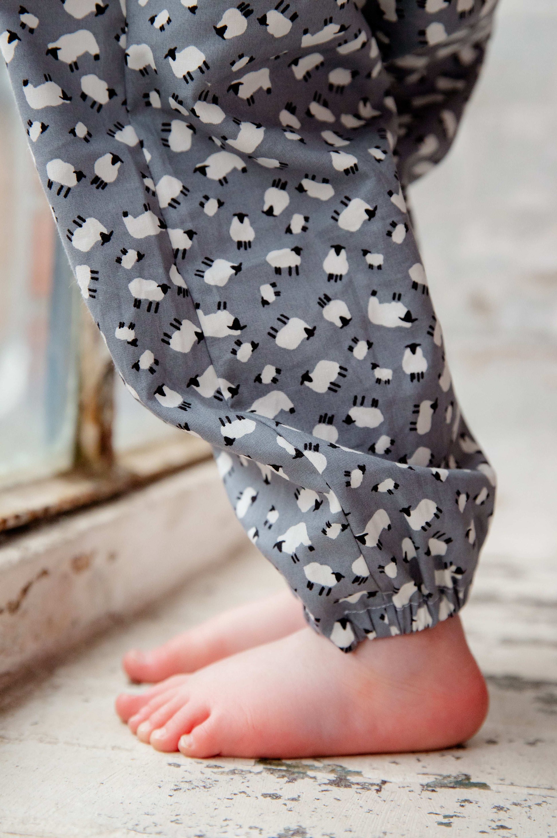 A young child wearing a handmade sheep romper. The romper has a delicate grey and white sheep print on soft, lightweight fabric. It has thin, shoulder straps and ruched neckline, relaxed, comfortable fit.