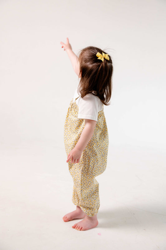 A young child wearing a handmade floral romper. The romper has a delicate yellow and white ditsy floral print on soft, lightweight fabric. It has thin, shoulder straps and ruched neckline, relaxed, comfortable fit.