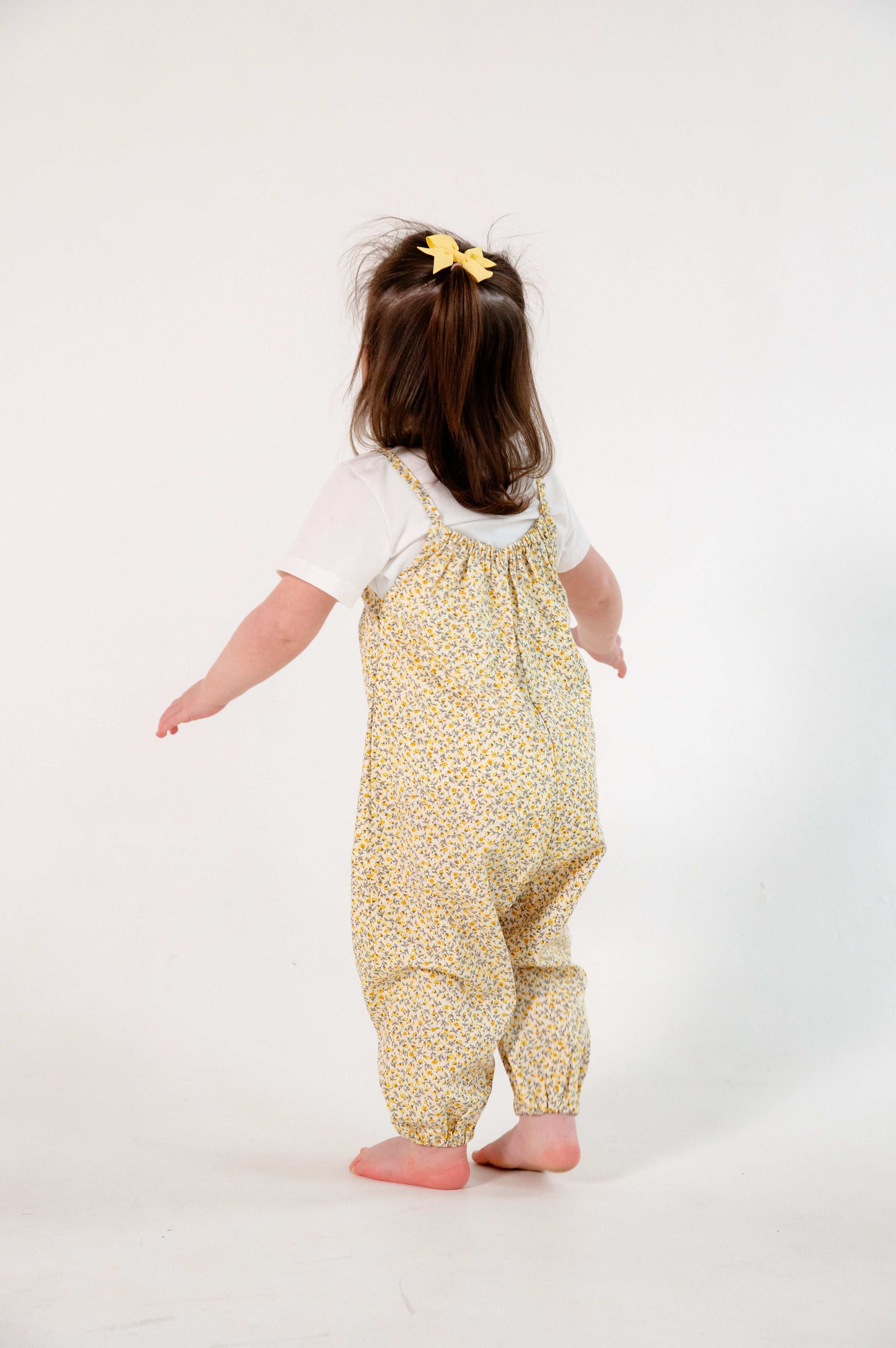 A young child wearing a handmade floral romper. The romper has a delicate yellow and white ditsy floral print on soft, lightweight fabric. It has thin, shoulder straps and ruched neckline, relaxed, comfortable fit.