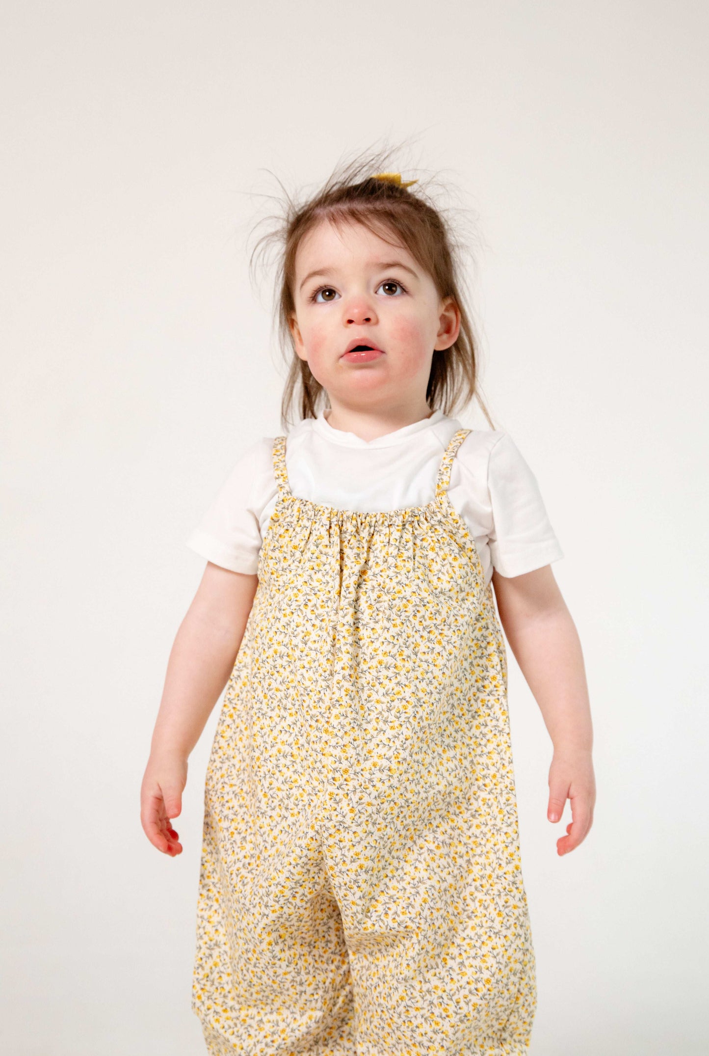 A young child wearing a handmade floral romper. The romper has a delicate yellow and white ditsy floral print on soft, lightweight fabric. It has thin, shoulder straps and ruched neckline, relaxed, comfortable fit.