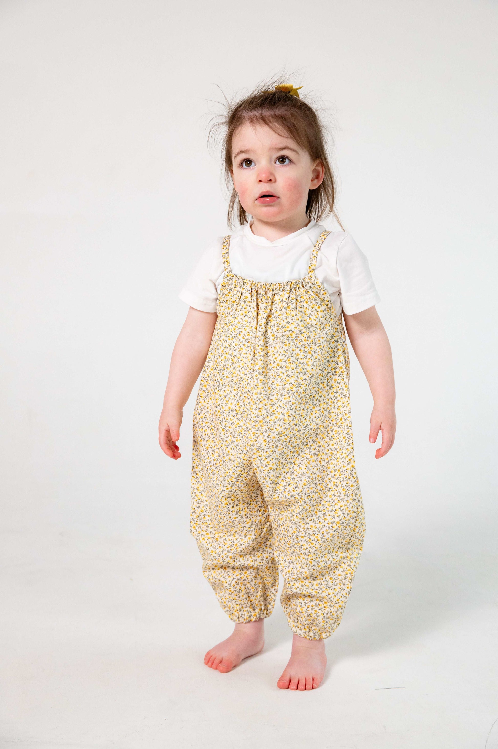 A young child wearing a handmade floral romper. The romper has a delicate yellow and white ditsy floral print on soft, lightweight fabric. It has thin, shoulder straps and ruched neckline, relaxed, comfortable fit.