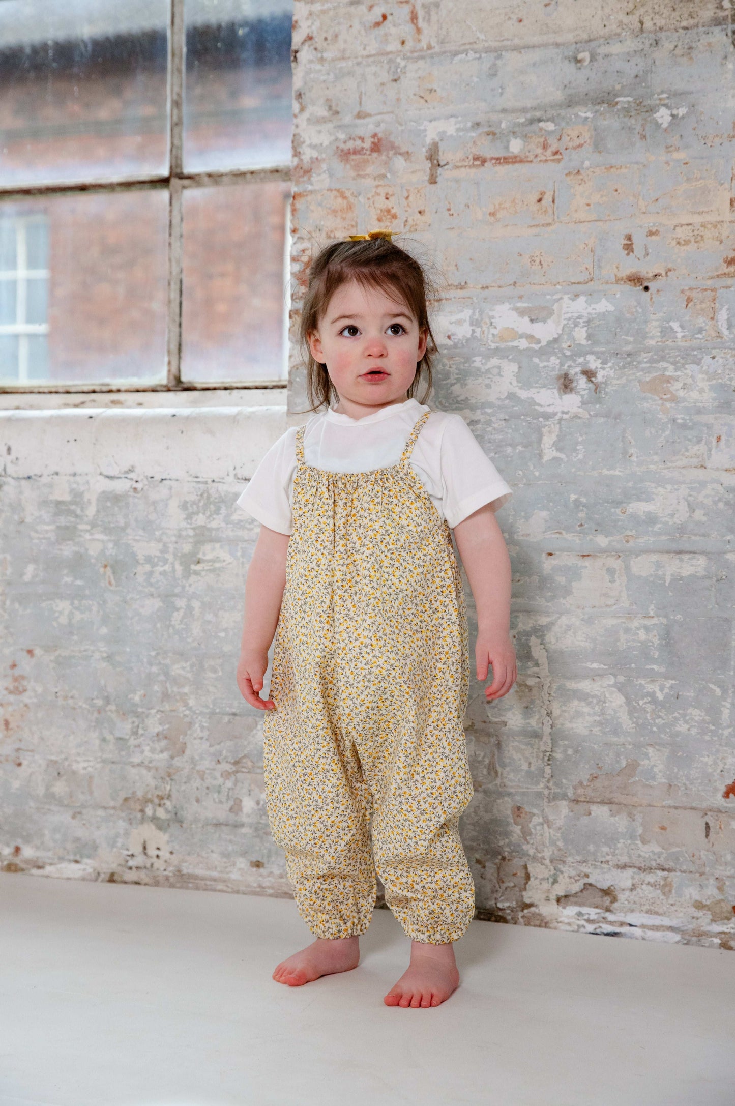A young child wearing a handmade floral romper. The romper has a delicate yellow and white ditsy floral print on soft, lightweight fabric. It has thin, shoulder straps and ruched neckline, relaxed, comfortable fit.
