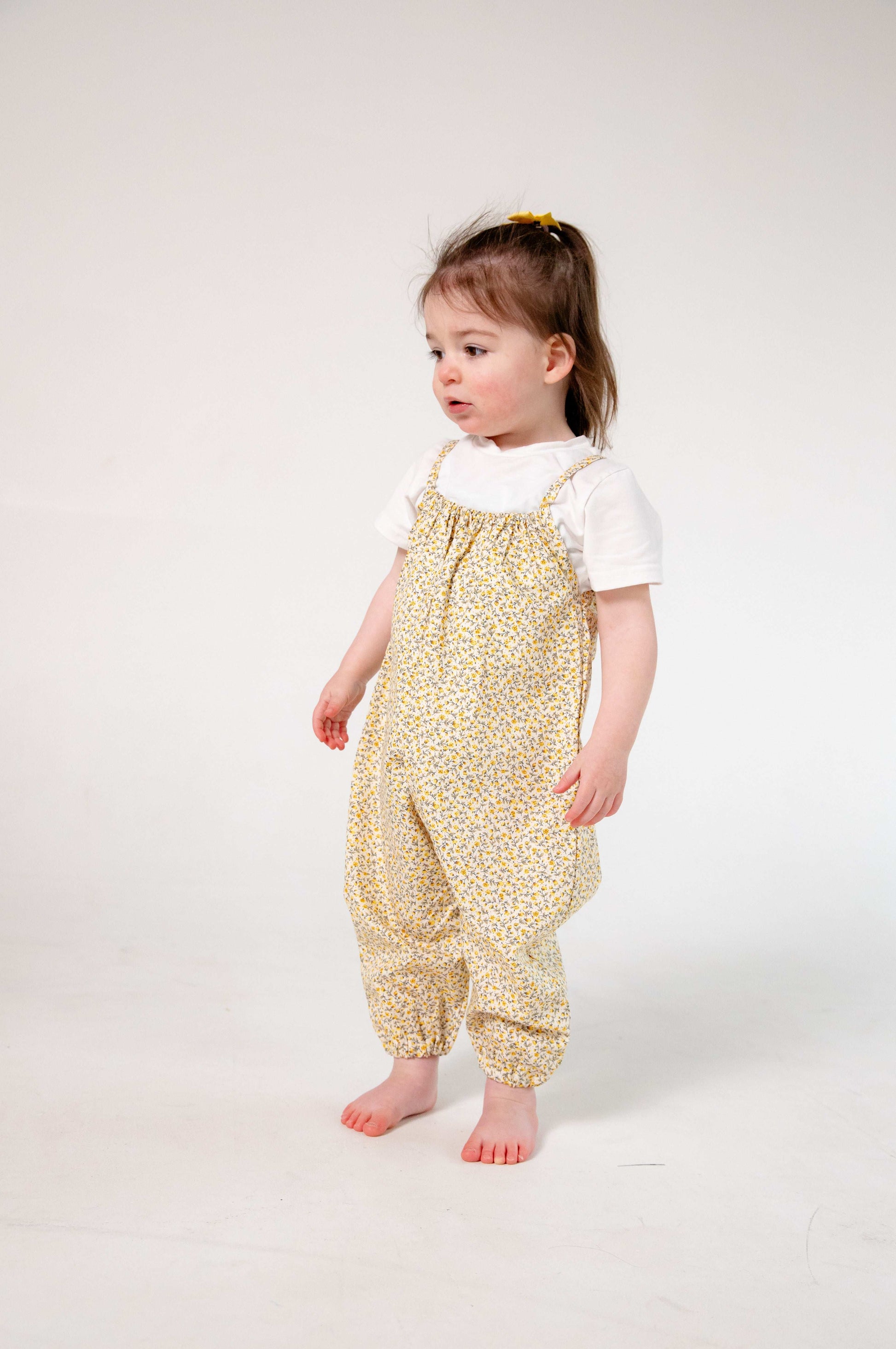 A young child wearing a handmade floral romper. The romper has a delicate yellow and white ditsy floral print on soft, lightweight fabric. It has thin, shoulder straps and ruched neckline, relaxed, comfortable fit.