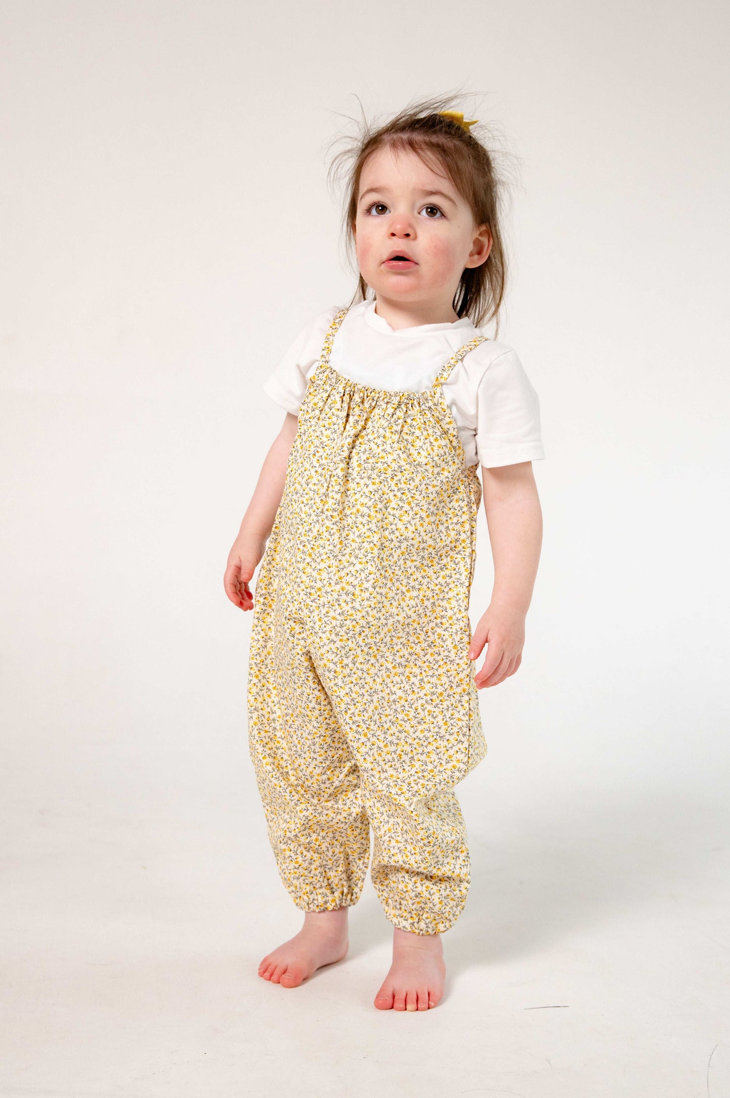 A young child wearing a handmade floral romper. The romper has a delicate yellow and white ditsy floral print on soft, lightweight fabric. It has thin, shoulder straps and ruched neckline, relaxed, comfortable fit.