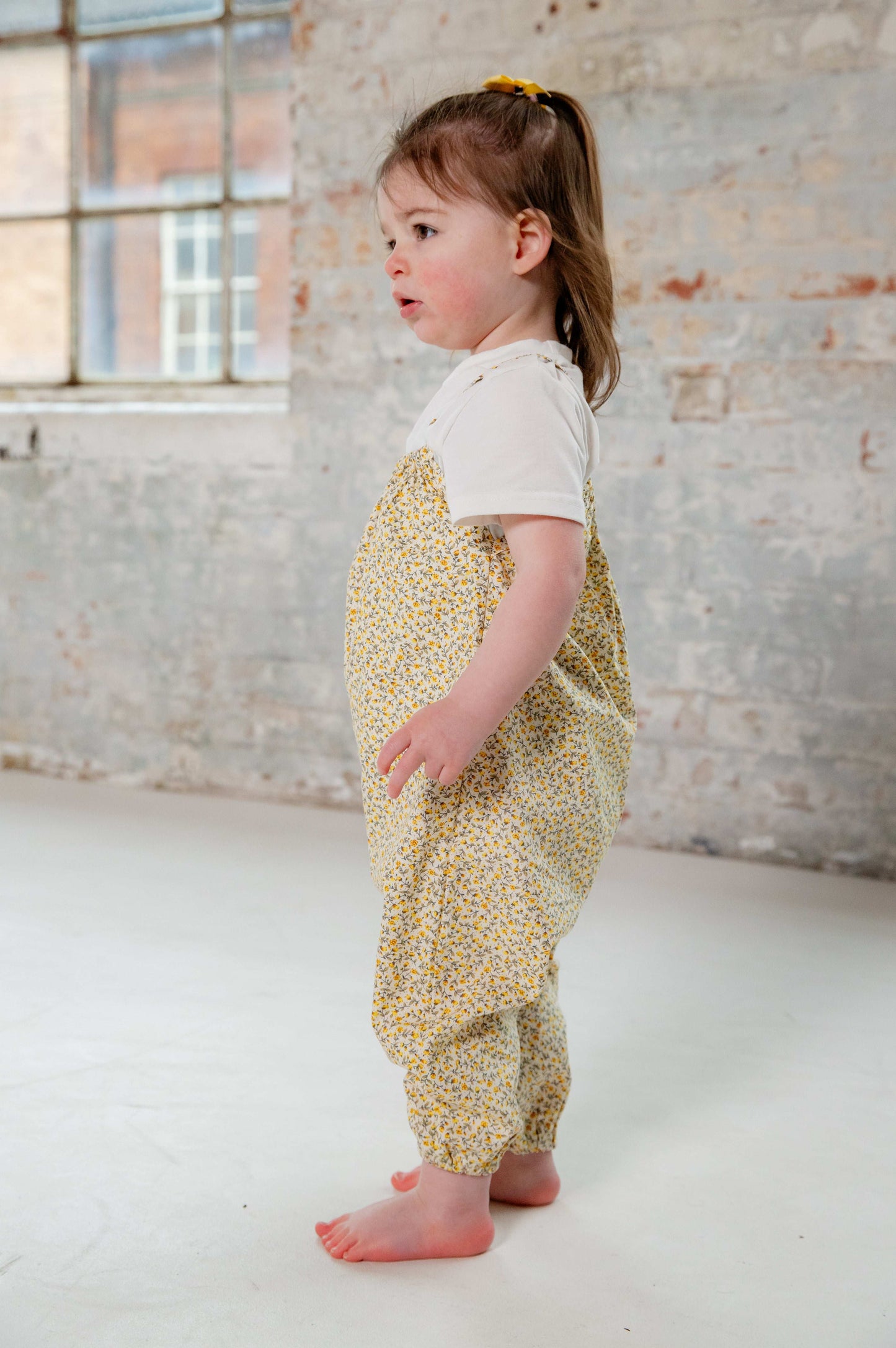 A young child wearing a handmade floral romper. The romper has a delicate yellow and white ditsy floral print on soft, lightweight fabric. It has thin, shoulder straps and ruched neckline, relaxed, comfortable fit.