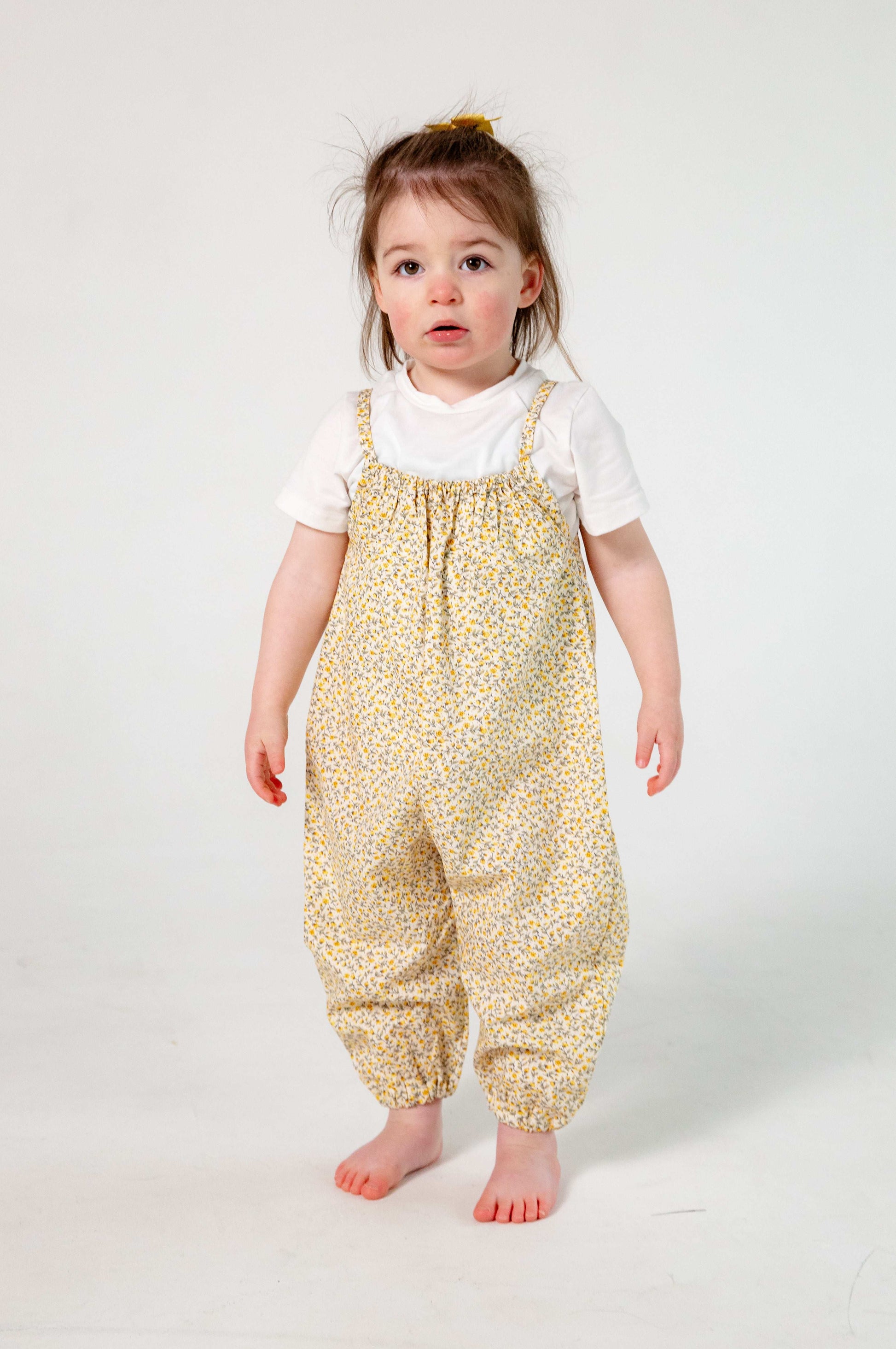 A young child wearing a handmade floral romper. The romper has a delicate yellow and white ditsy floral print on soft, lightweight fabric. It has thin, shoulder straps and ruched neckline, relaxed, comfortable fit.