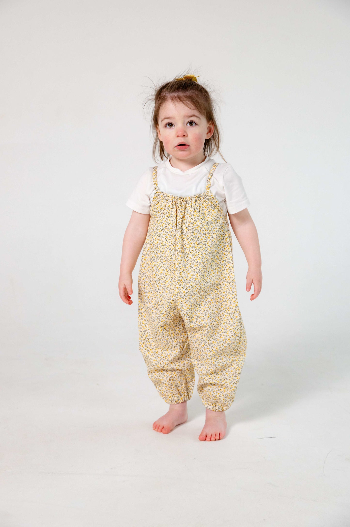 A young child wearing a handmade floral romper. The romper has a delicate yellow and white ditsy floral print on soft, lightweight fabric. It has thin, shoulder straps and ruched neckline, relaxed, comfortable fit.
