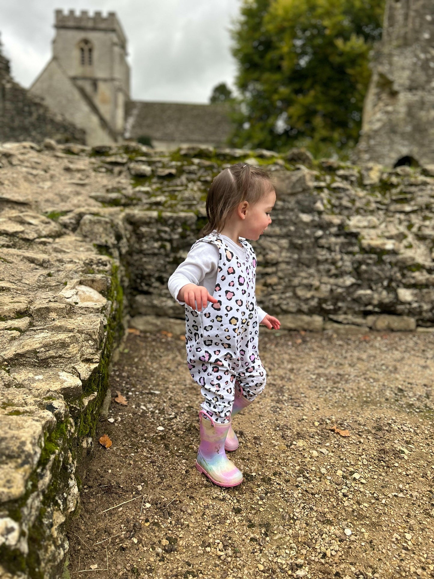 Photo of a toddler running in a romper and wellies, handmade in Witney Oxfordshire and photographed at Minster Lovell Ruins. The romper is white with pastel rainbow leopard spot, stretchy and fun.