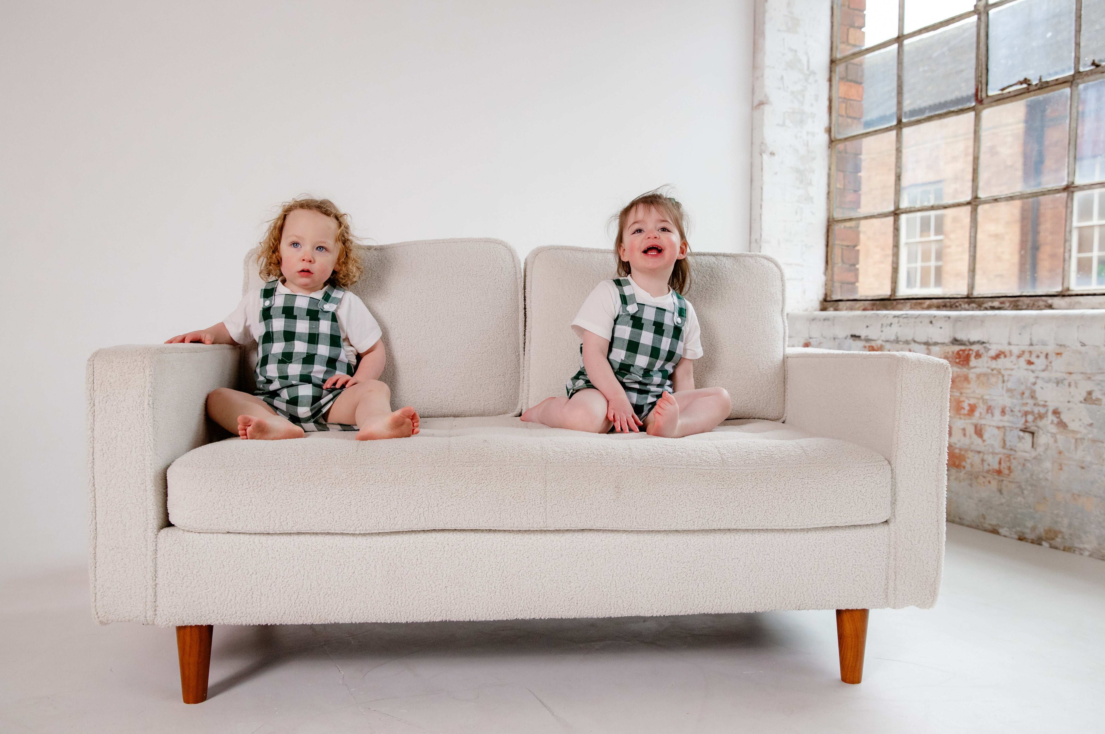 A toddler wearing a handmade green and white gingham romper crafted from cotton fabric. The romper features, a front pocket, and a loose comfortable fit. The outfit is a timeless, classic design, perfect for all occasions.