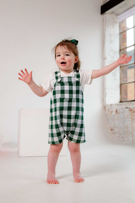 A toddler wearing a handmade green and white gingham romper crafted from cotton fabric. The romper features, a front pocket, and a loose comfortable fit. The outfit is a timeless, classic design, perfect for all occasions.