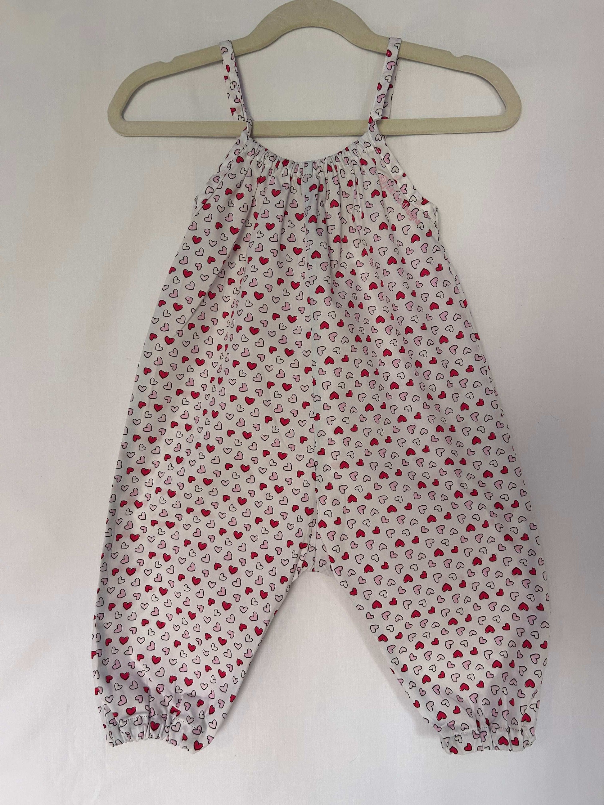 A young child wearing a handmade heart print romper. The romper has a bold heart print on soft, lightweight fabric. It has thin, shoulder straps and ruched neckline, relaxed, comfortable fit. 