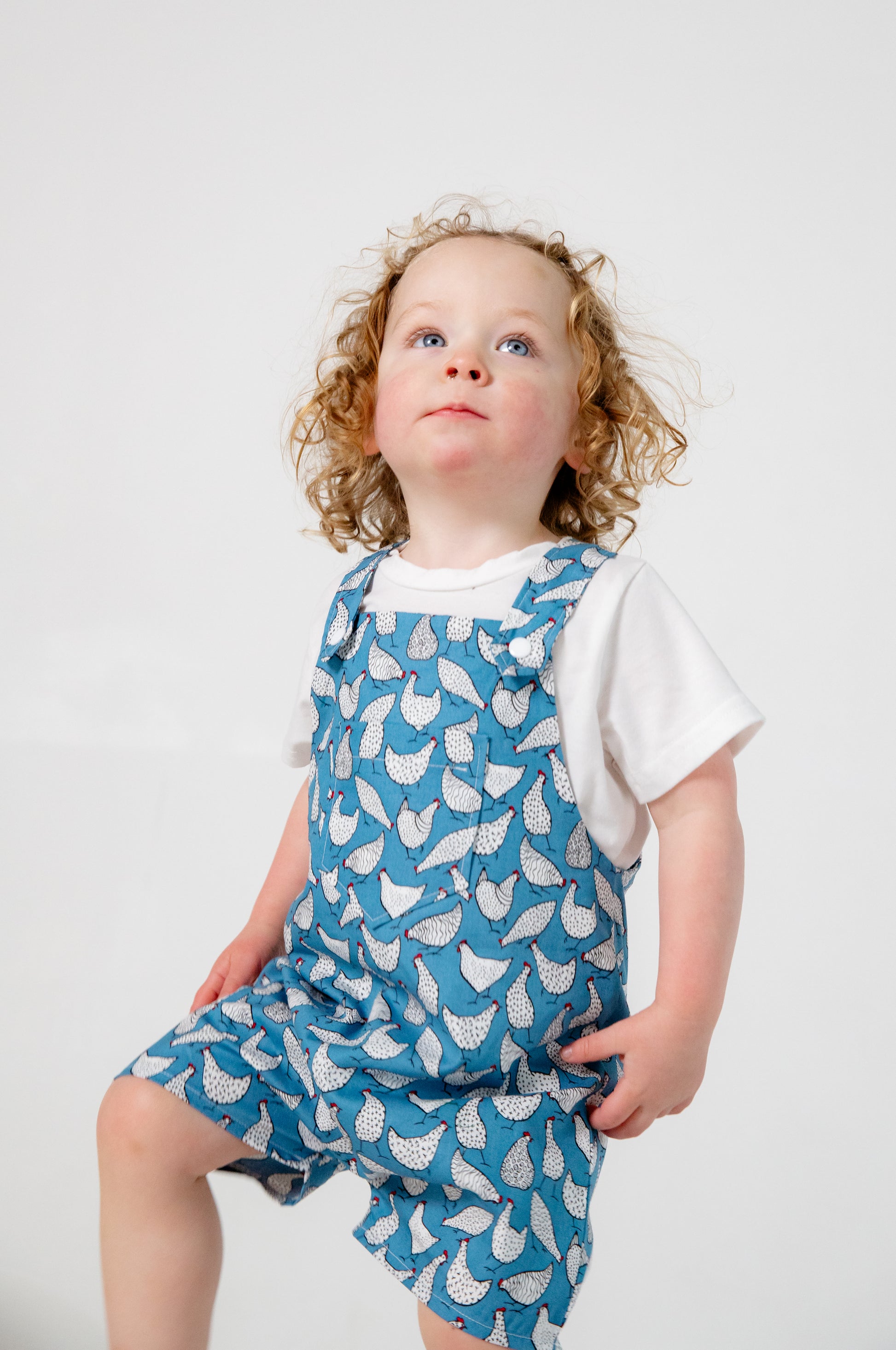 A young child wears handmade blue overalls featuring a playful white chicken pattern, crafted in Oxfordshire. The overalls are paired with a simple white t-shirt. The high-quality fabric and thoughtful design highlight the craftsmanship of the garment. The child, with curly blonde hair and fair skin, stands against a plain white background, looking upward with a curious expression.