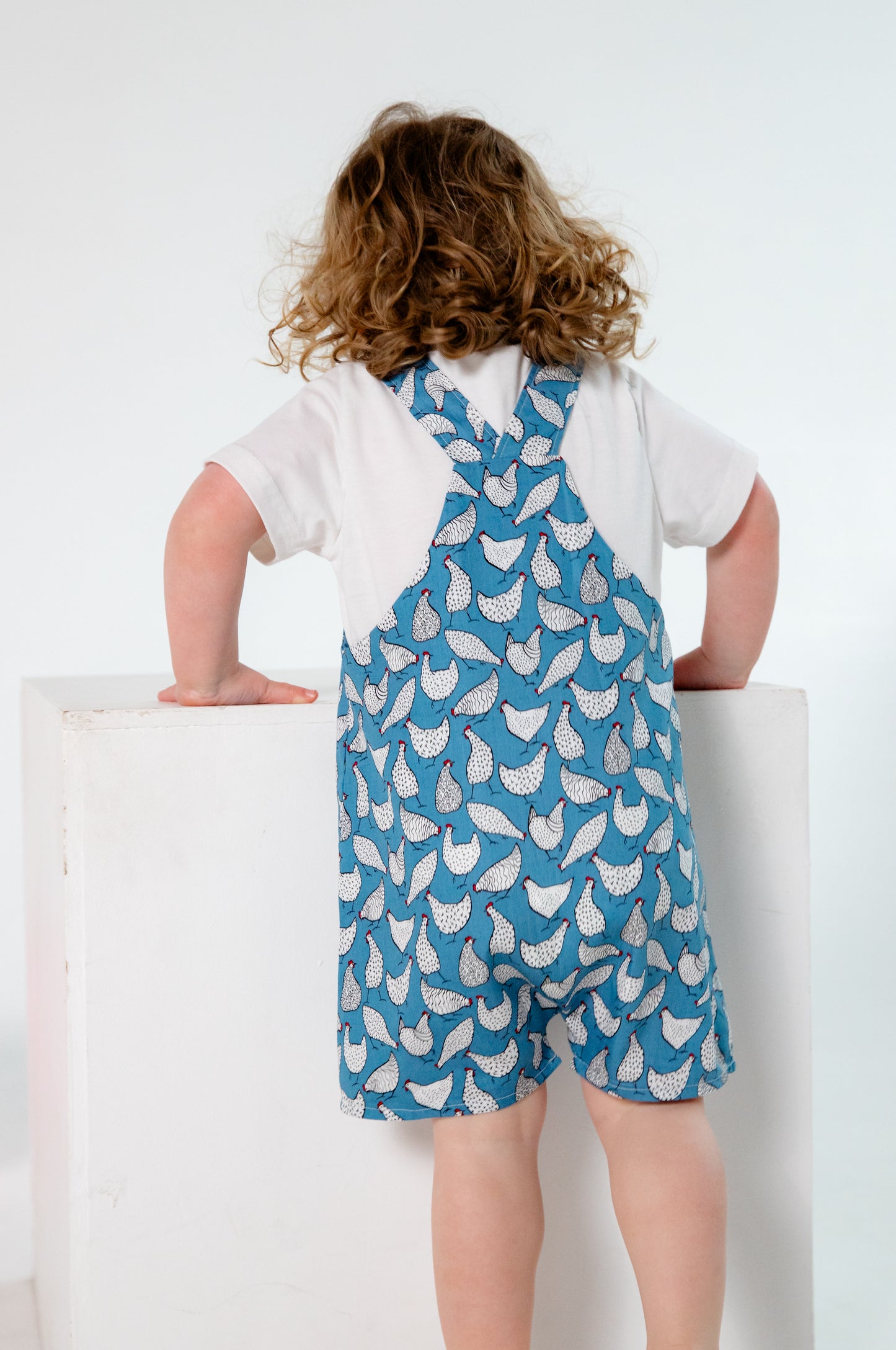 A young child wears handmade blue overalls featuring a playful white chicken pattern, crafted in Oxfordshire. The overalls are paired with a simple white t-shirt. The high-quality fabric and thoughtful design highlight the craftsmanship of the garment. The child, with curly blonde hair and fair skin, stands against a plain white background, looking upward with a curious expression.