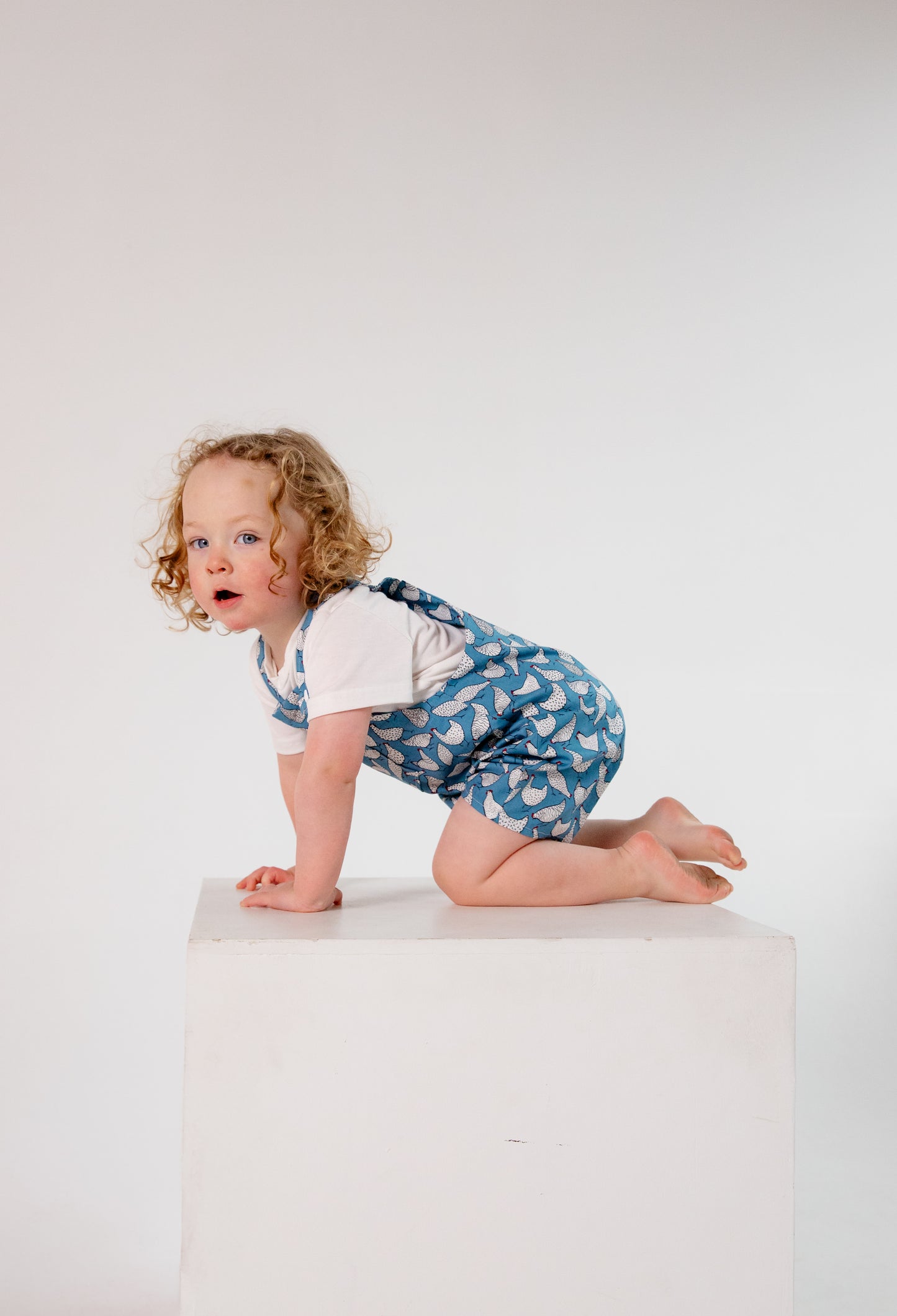 A young child wears handmade blue overalls featuring a playful white chicken pattern, crafted in Oxfordshire. The overalls are paired with a simple white t-shirt. The high-quality fabric and thoughtful design highlight the craftsmanship of the garment. The child, with curly blonde hair and fair skin, stands against a plain white background, looking upward with a curious expression.