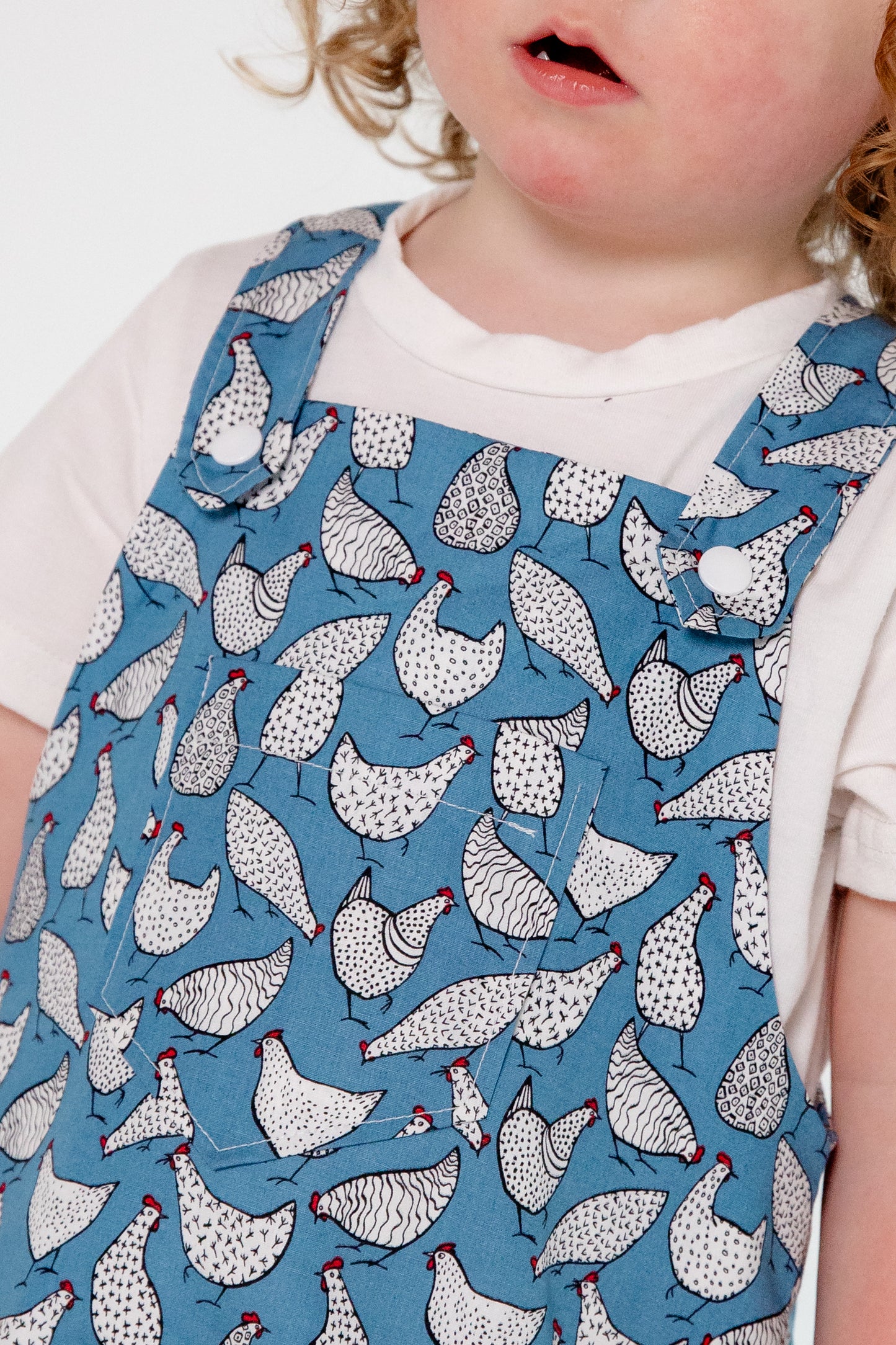 A young child wears handmade blue overalls featuring a playful white chicken pattern, crafted in Oxfordshire. The overalls are paired with a simple white t-shirt. The high-quality fabric and thoughtful design highlight the craftsmanship of the garment. The child, with curly blonde hair and fair skin, stands against a plain white background, looking upward with a curious expression.