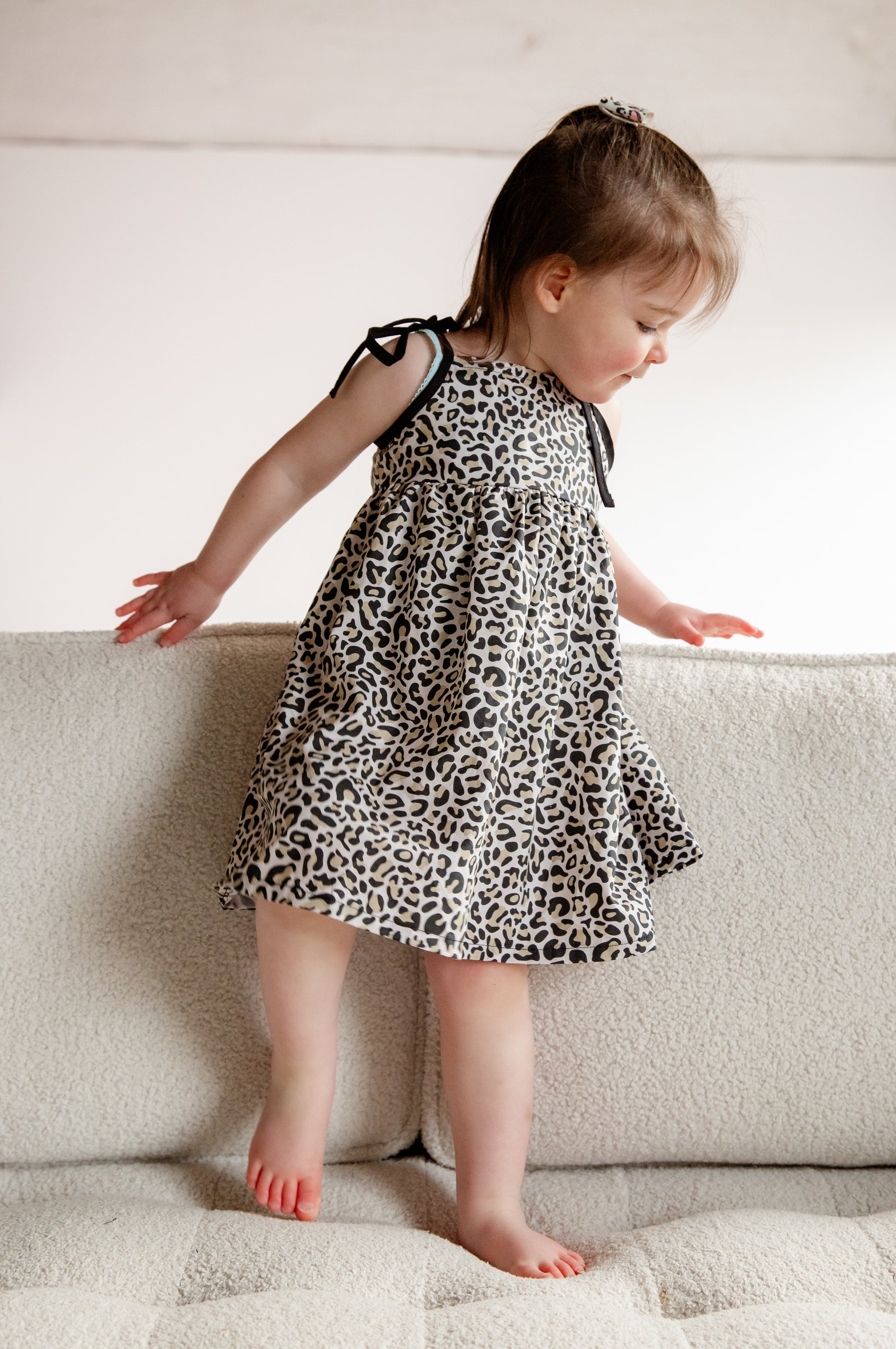 A young child wearing a handmade dress from Oxfordshire featuring a black and white leopard print pattern. The dress has a sleeveless design with delicate black ribbon ties on the shoulders, a gathered waistline, and a flowing skirt. The fabric has a soft yet structured appearance, complementing the child’s playful movement. The child is barefoot, standing on a textured cream-colored couch, adding to the cozy and natural feel of the image.