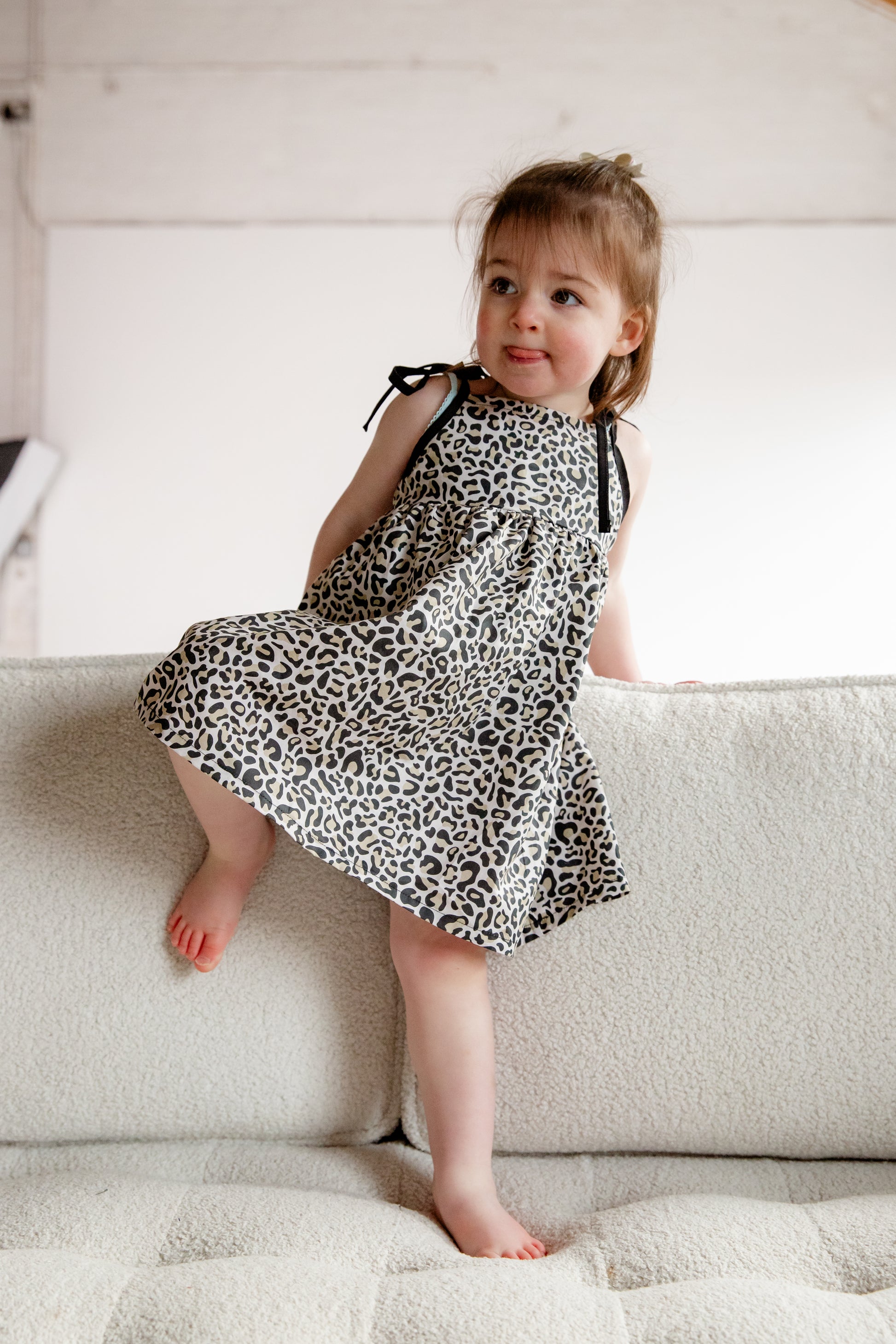 A young child wearing a handmade dress from Oxfordshire featuring a black and white leopard print pattern. The dress has a sleeveless design with delicate black ribbon ties on the shoulders, a gathered waistline, and a flowing skirt. The fabric has a soft yet structured appearance, complementing the child’s playful movement. The child is barefoot, standing on a textured cream-colored couch, adding to the cozy and natural feel of the image.