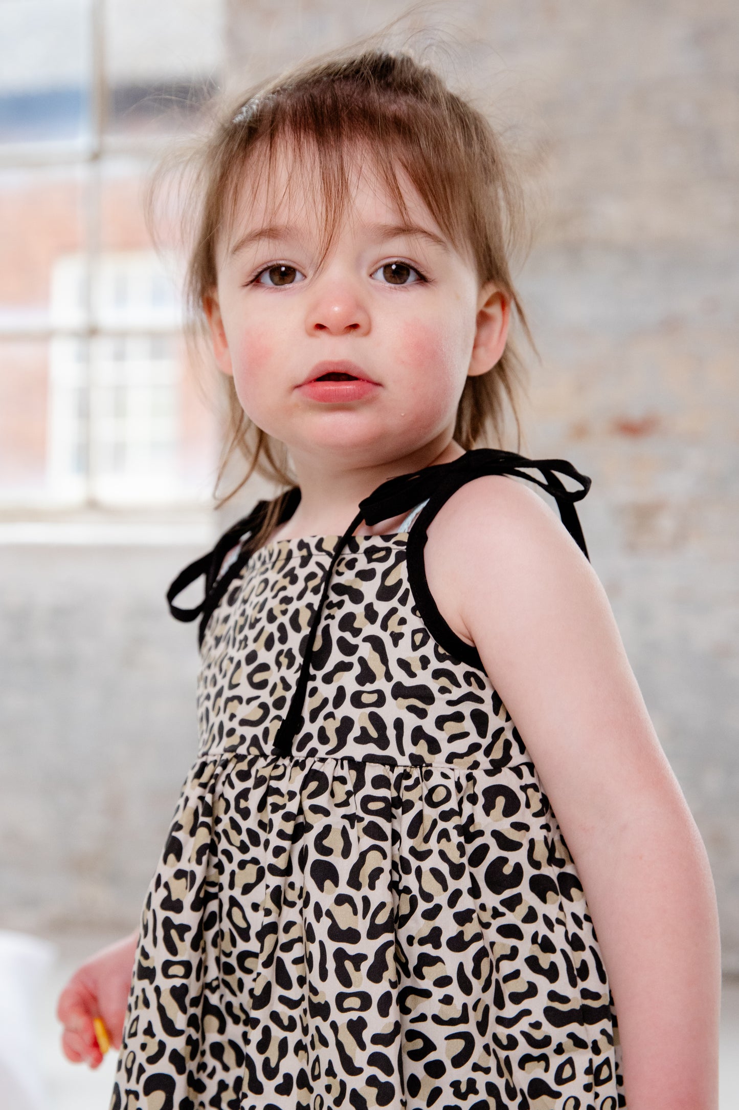 A young child wearing a handmade dress from Oxfordshire featuring a black and white leopard print pattern. The dress has a sleeveless design with delicate black ribbon ties on the shoulders, a gathered waistline, and a flowing skirt. The fabric has a soft yet structured appearance, complementing the child’s playful movement. The child is barefoot, standing on a textured cream-colored couch, adding to the cozy and natural feel of the image.